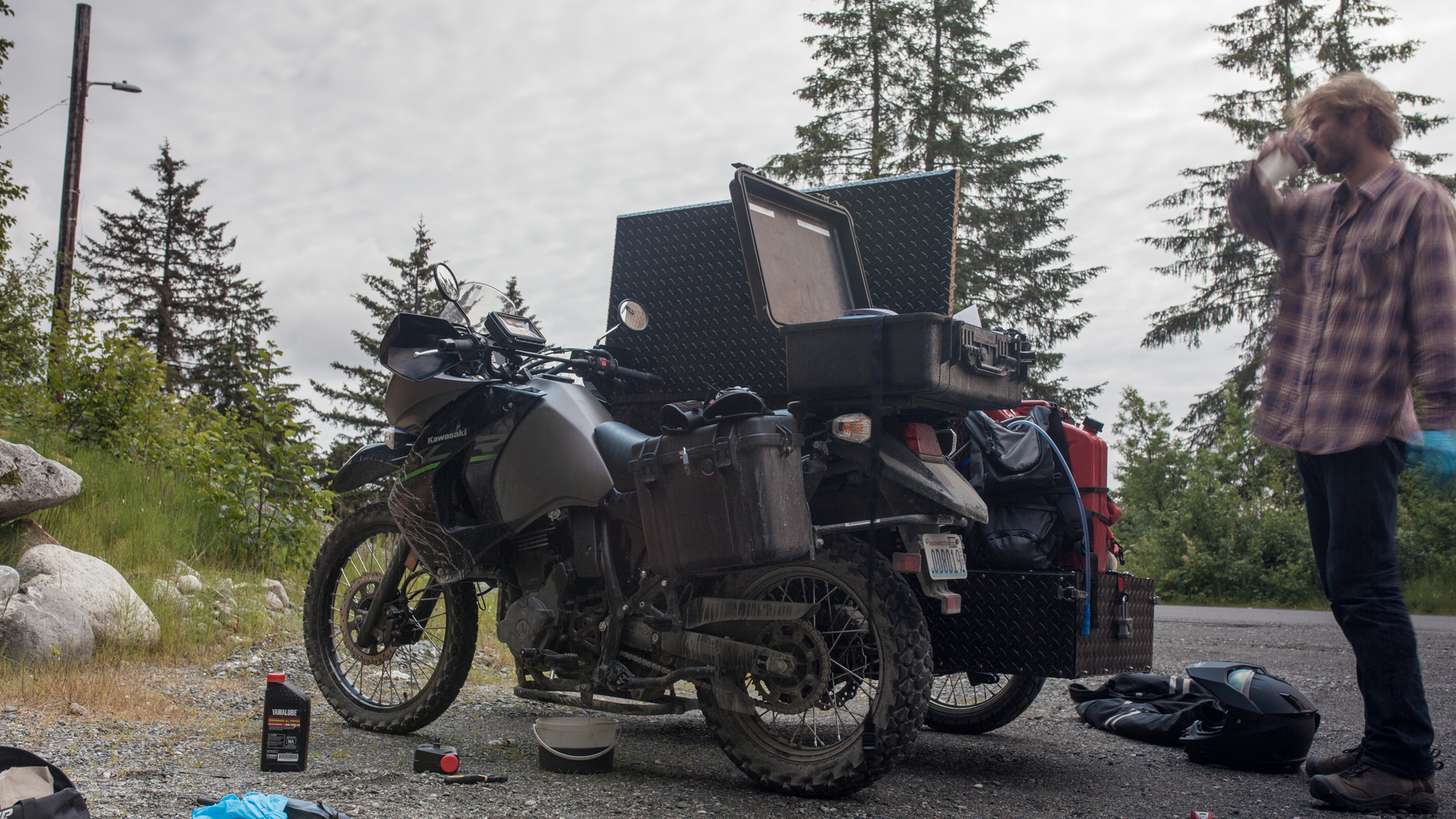 A large, rugged adventure motorcycle parked on a gravel road in a forest setting, with a person standing nearby.