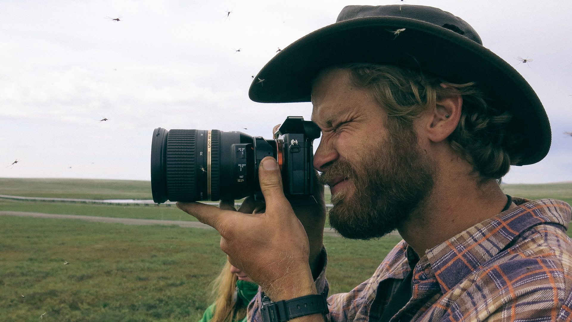 A bearded man in a cowboy hat using a large camera to take a photograph outdoors.