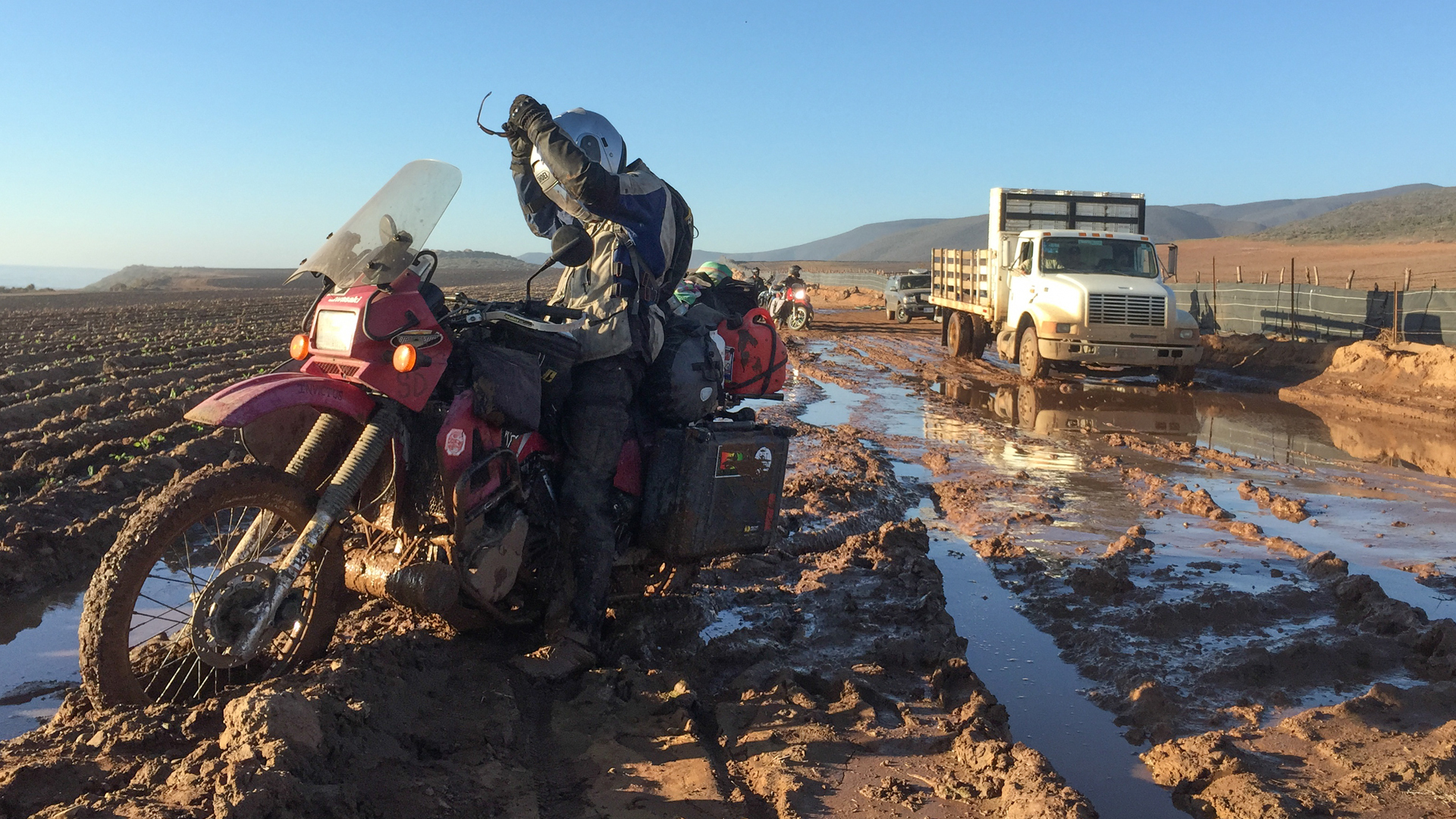 Muddy motorcycle in remote desert, person riding across flooded terrain.