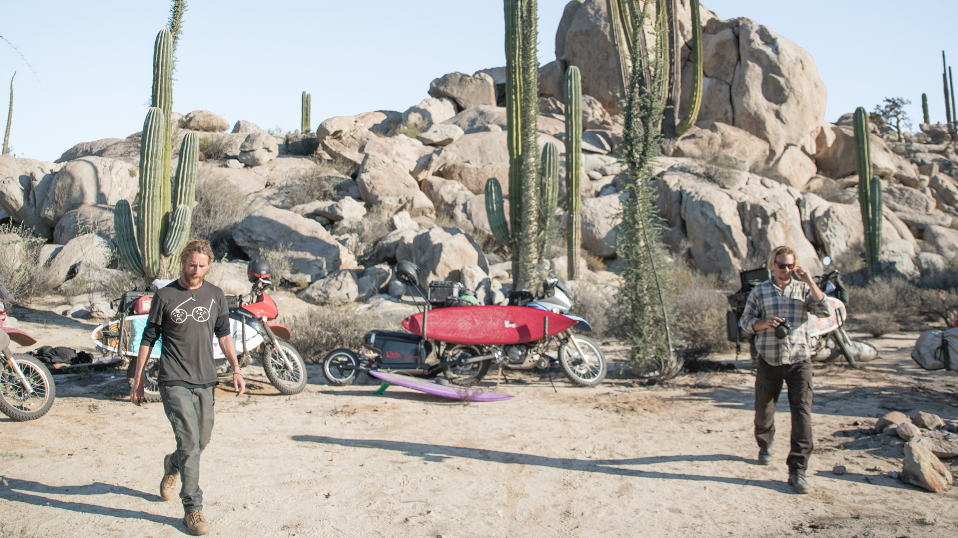 Arid desert landscape with rocky hills, cacti, and two individuals carrying motorcycles.