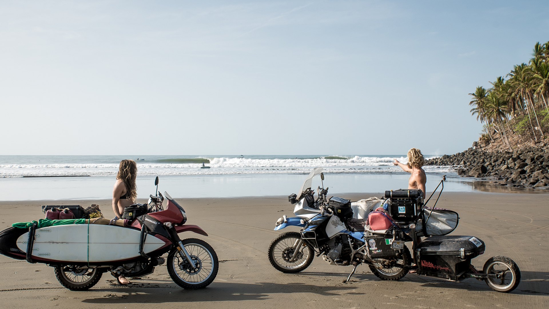 Two motorbikes parked on a beach, one with a surfboard attached, beside palm trees and the ocean.
