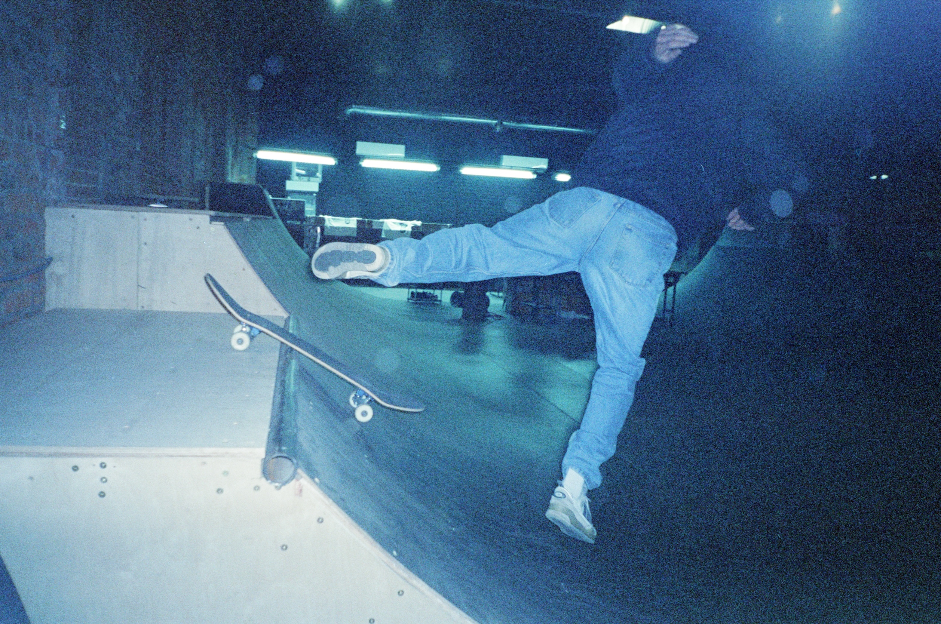 Dark indoor skate park with a person riding a skateboard down a curved ramp, wearing protective gear.