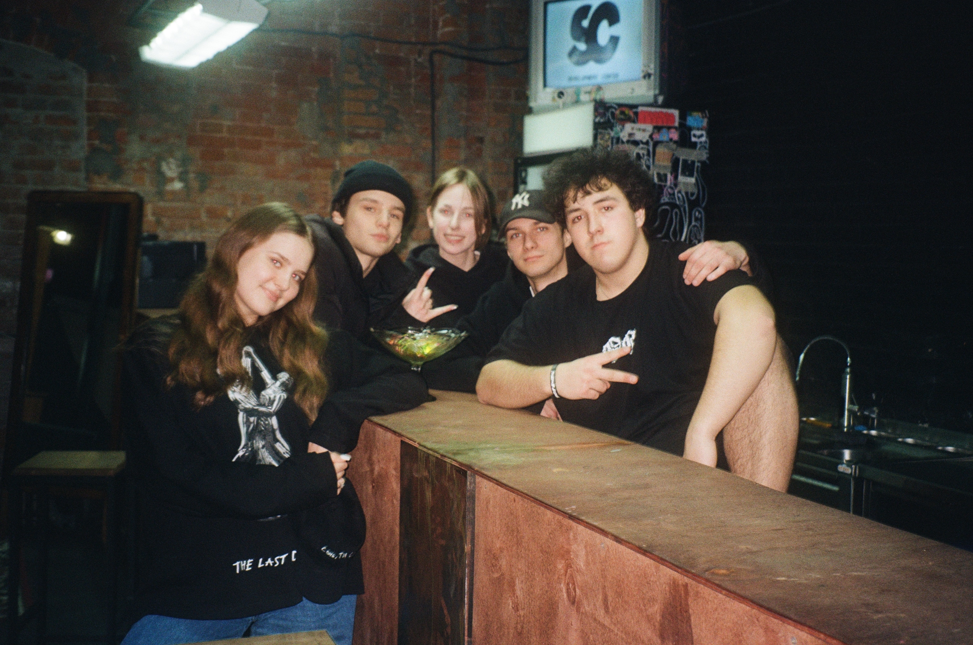 Group of 5 people, including 3 men and 2 women, posing in a dimly lit room with assorted items and equipment visible.