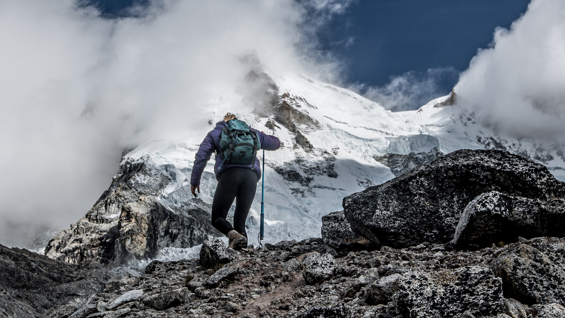 Hiker on a rocky path, surrounded by snow-capped mountains and cloudy skies.