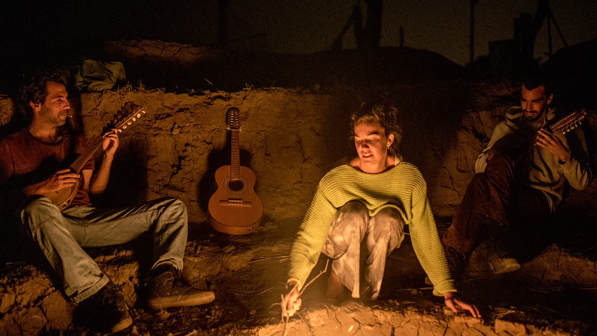 Men playing guitars and singing around a fire at night.