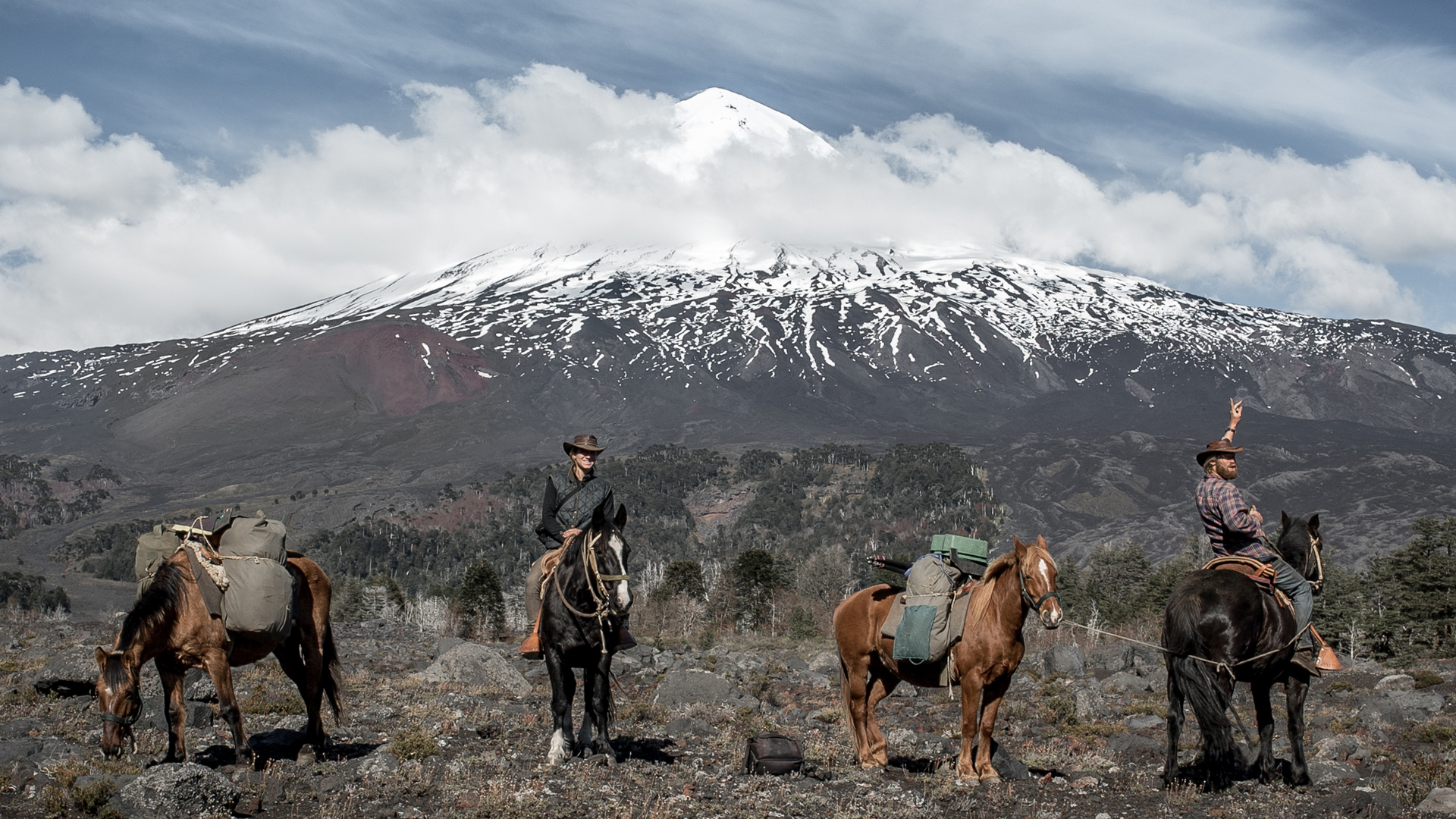 Snowy mountain peak, cloudy sky, people on horseback in a field