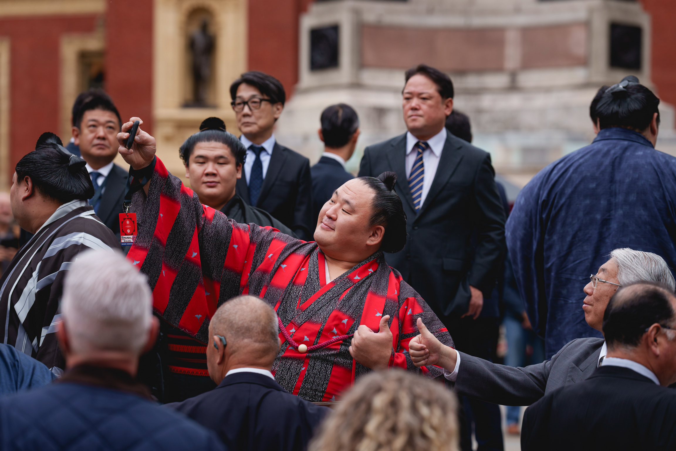 Large sumo wrestler in red ceremonial robe surrounded by crowd of people in dark suits at formal outdoor event.