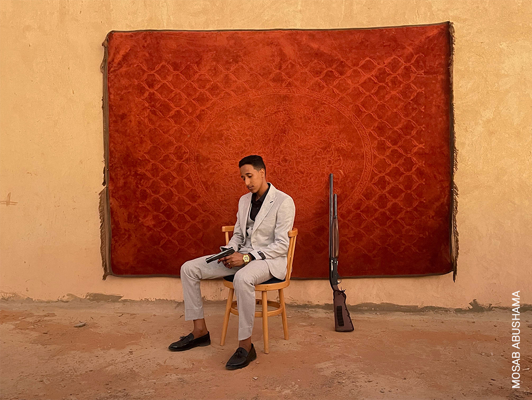 A man in a white suit sits on a chair in front of a red patterned wall, holding a rifle.