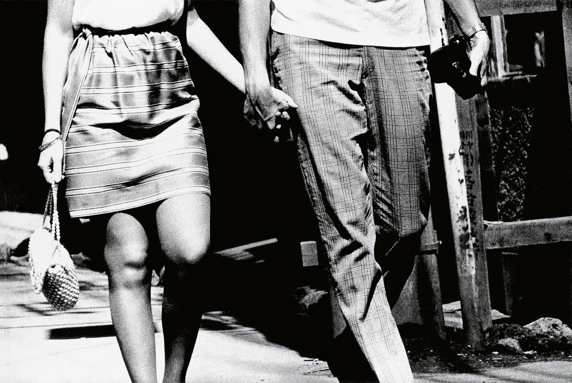 Black and white image of couple holding hands walking, woman in striped skirt and white top, man in checked trousers and shirt.