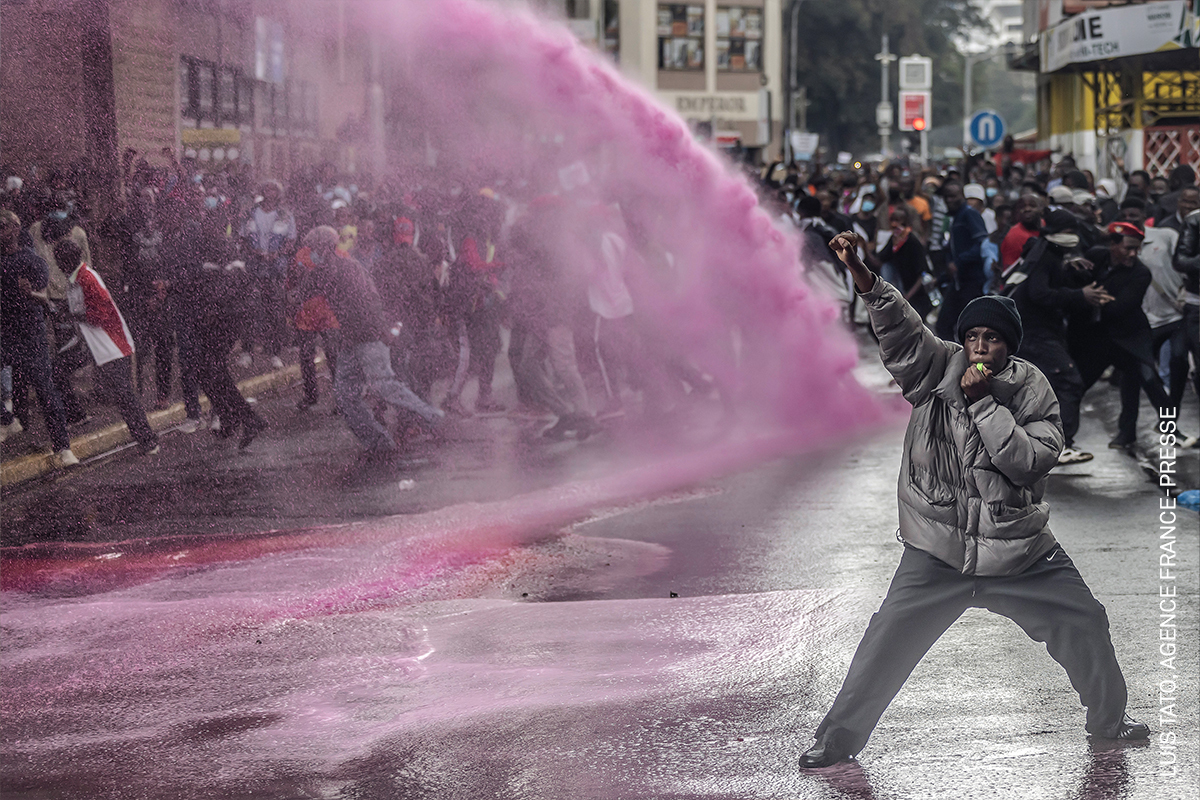 Crowd of protesters in street, police using water cannon, single individual in foreground shielding themselves, pink liquid spraying through the air.