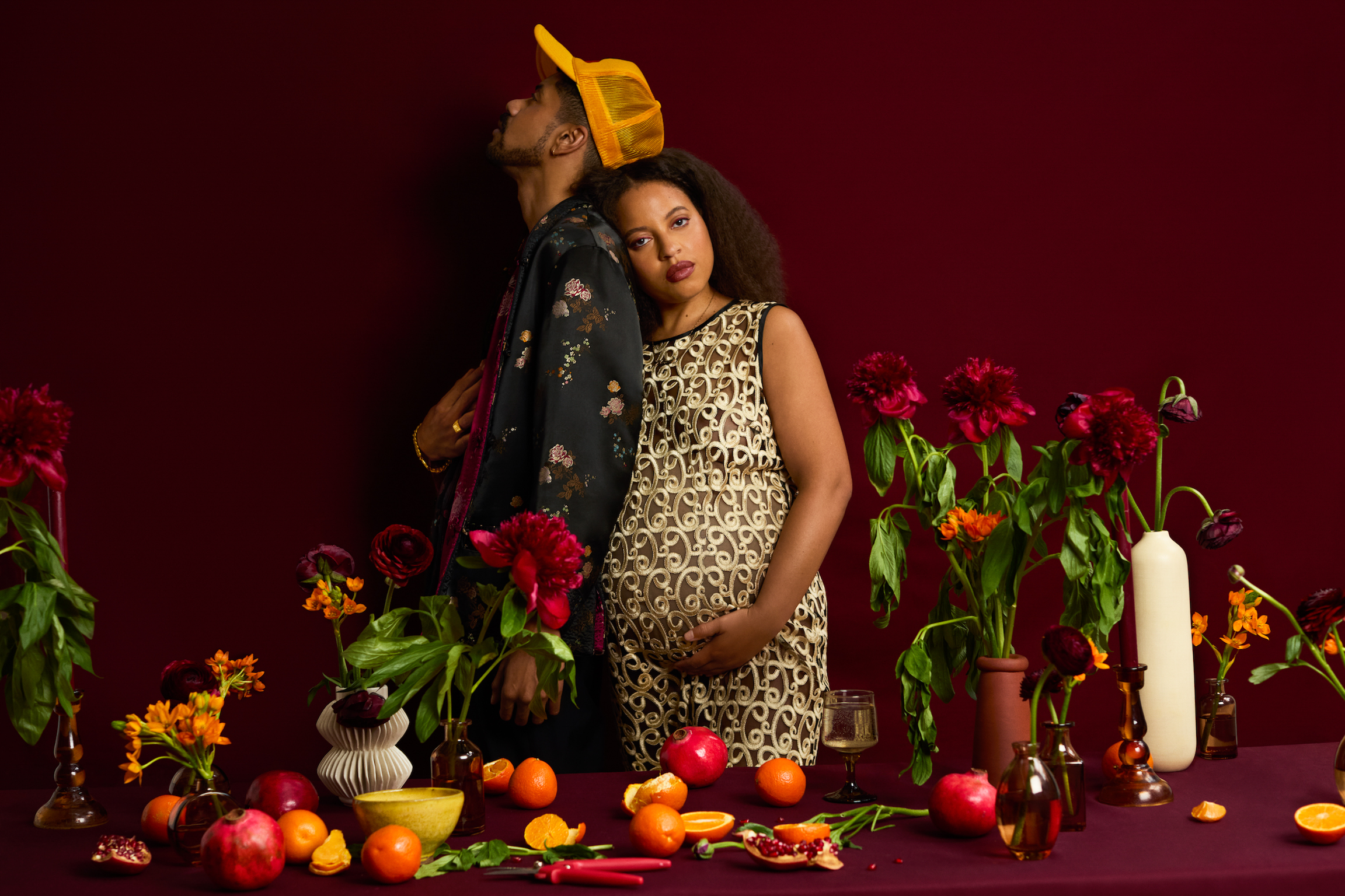 Man in yellow cap kisses pregnant woman in leopard print dress surrounded by flowers, fruit and vegetables on dark red background.