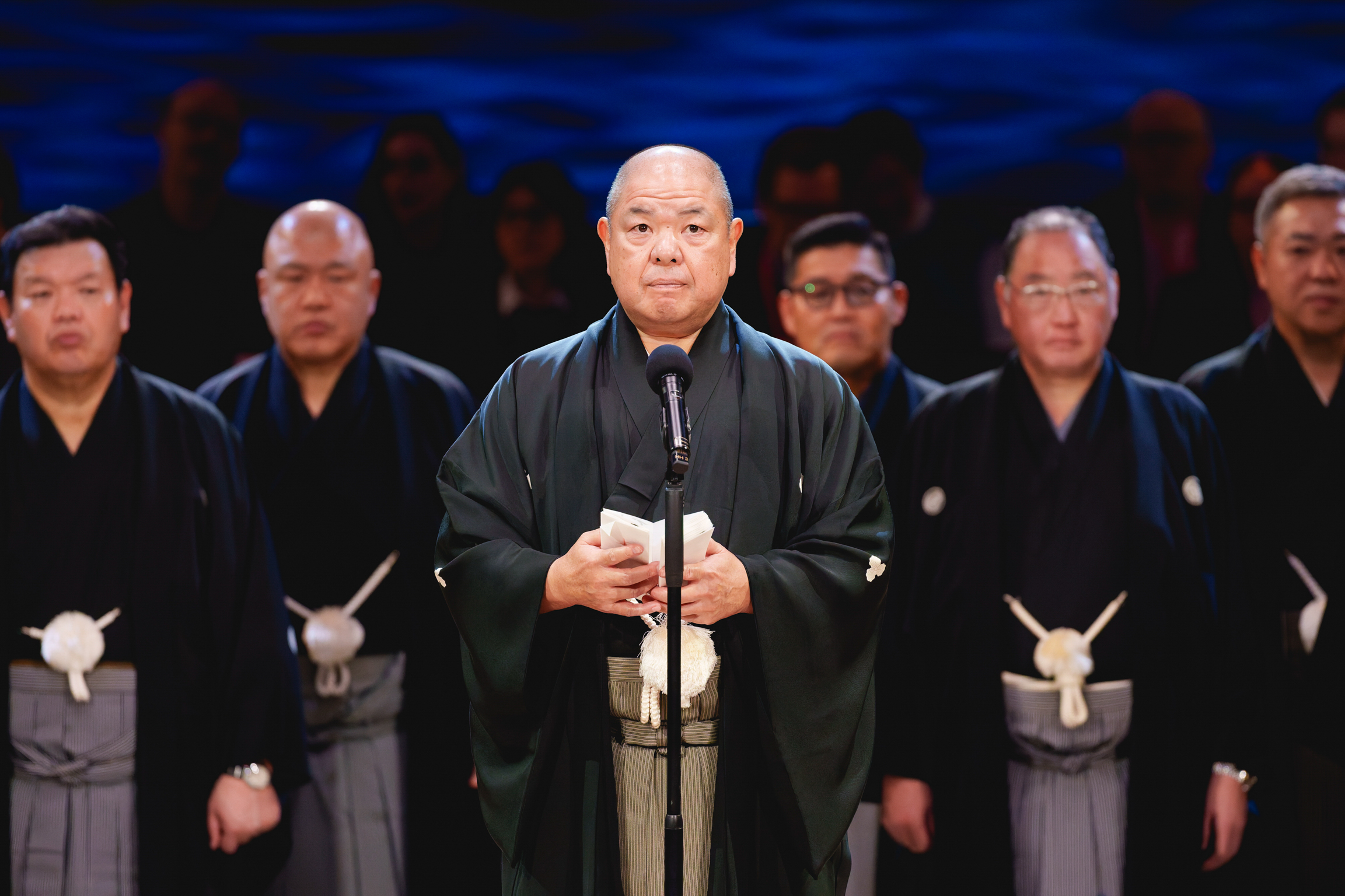 Bald man in black robes holding sword, standing before microphone with similarly dressed men behind him against blue backdrop.