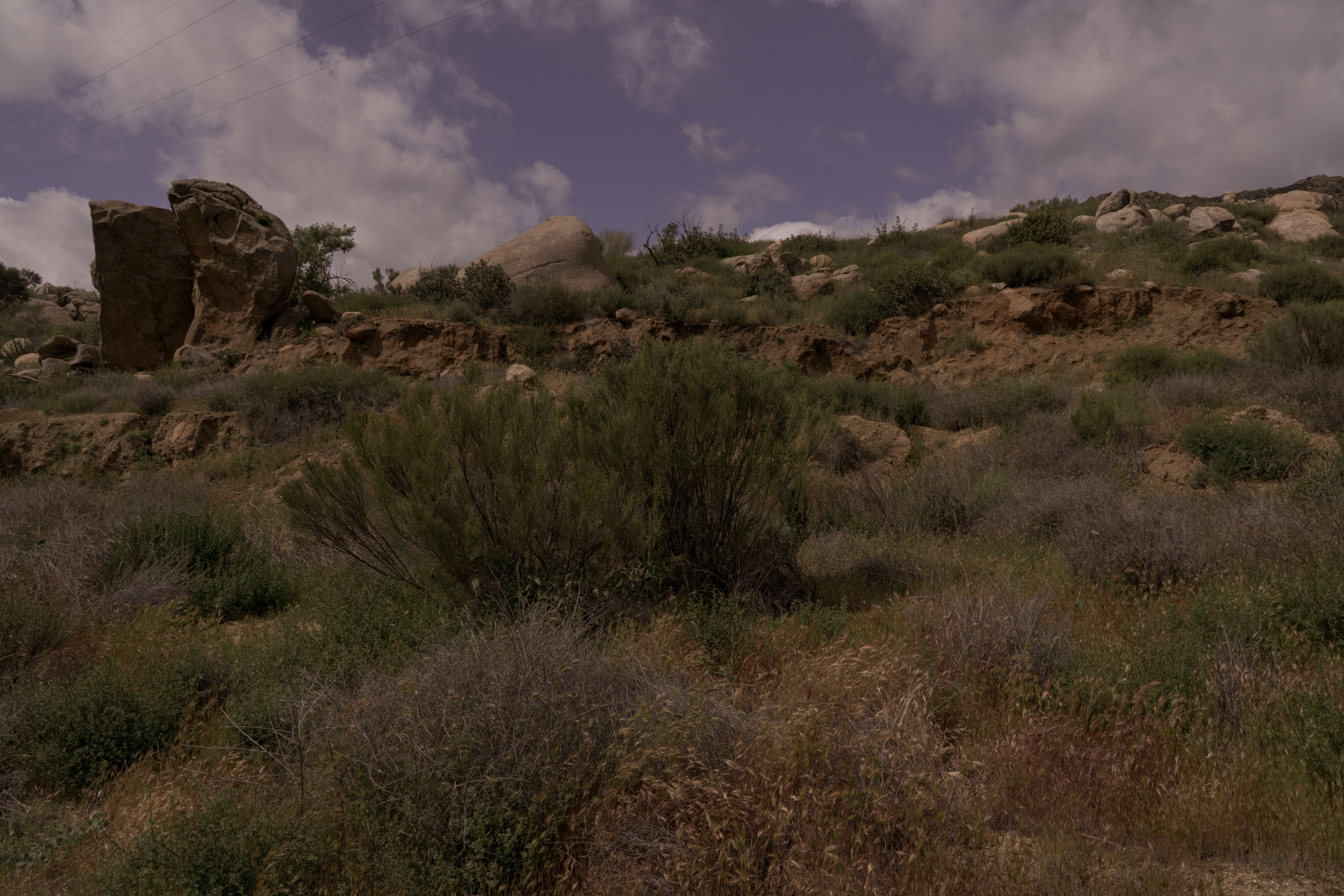 Rugged desert landscape with rocky outcrops, scrubby vegetation, and a cloudy sky.