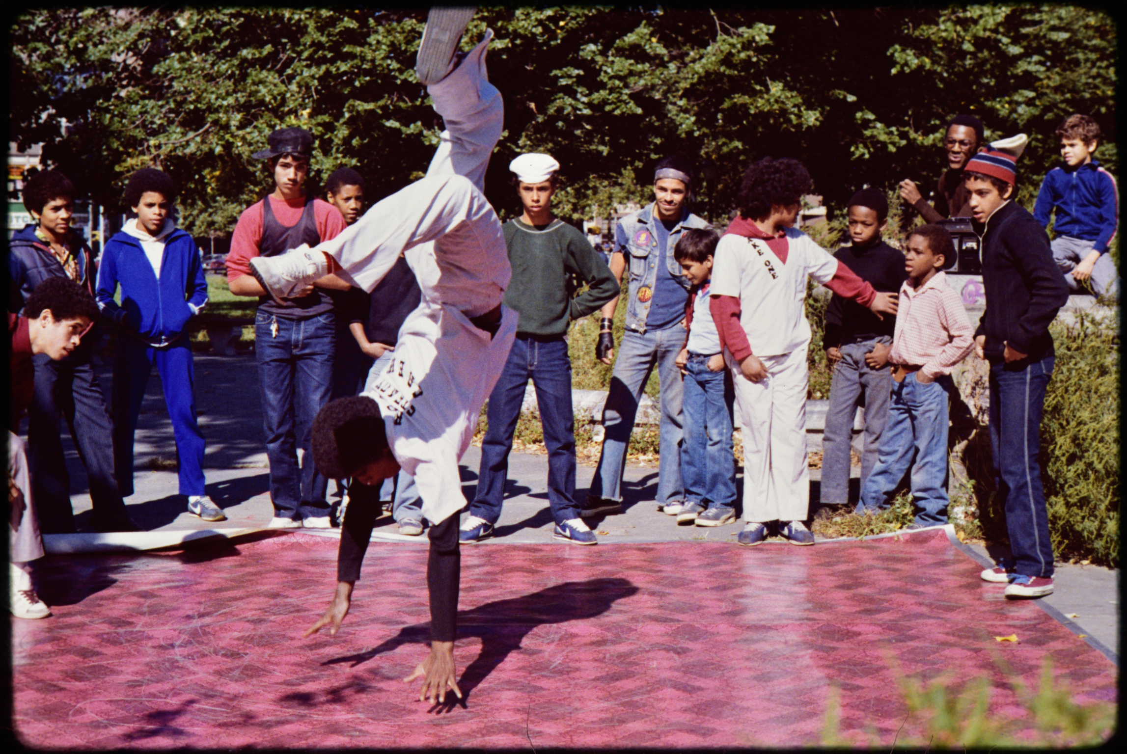 Breakdancer performing freeze move on cardboard mat surrounded by crowd of spectators in park setting with trees in background.