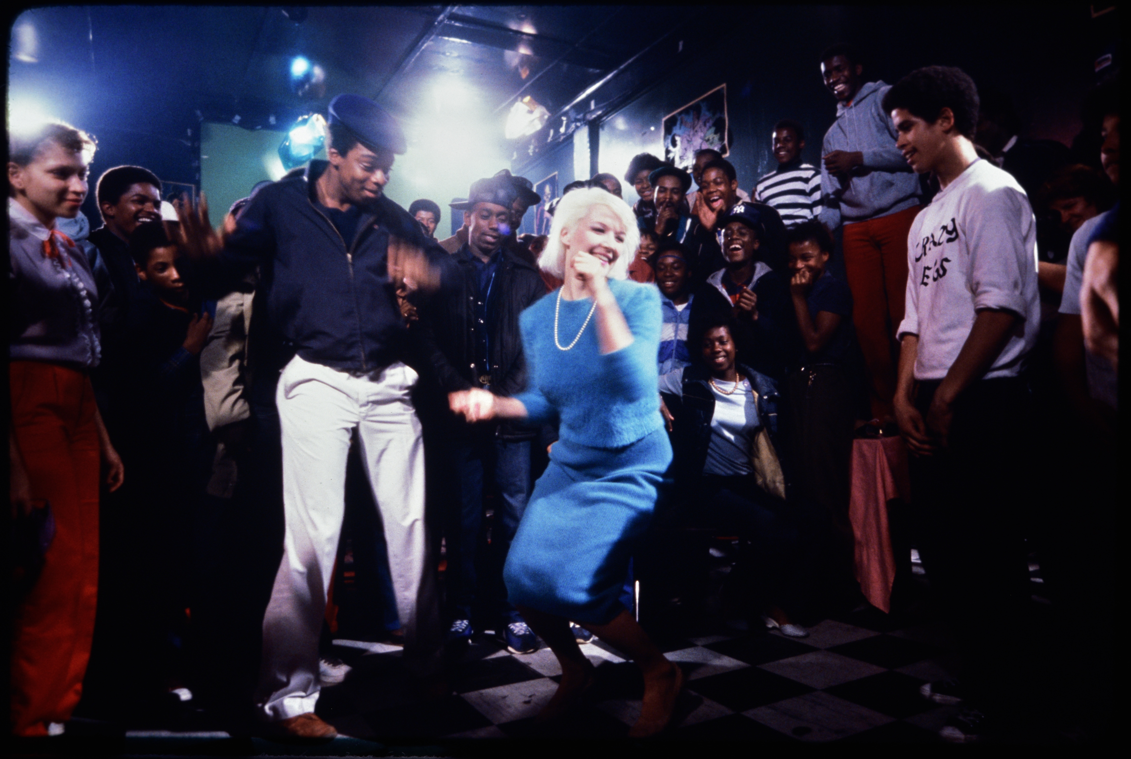 Blonde woman in blue dress dancing with crowd in dimly lit venue with stage lighting and gathered audience.