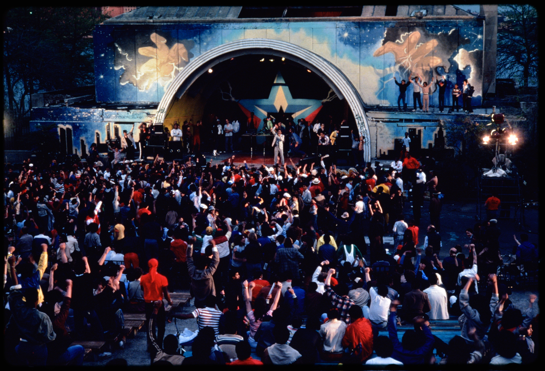 Large crowd gathered before ornate arched stage with blue backdrop featuring golden winged figures and starry night sky design.