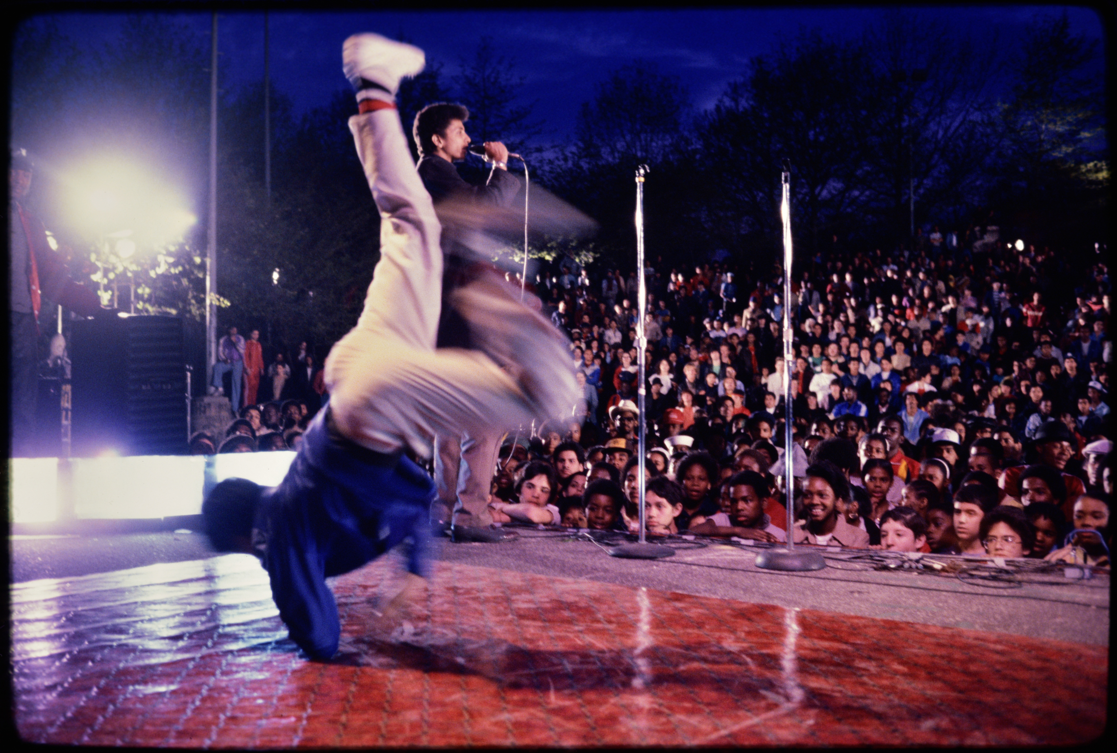 Performer in white shirt and dark trousers mid-jump on illuminated stage before large seated crowd at night-time outdoor venue.