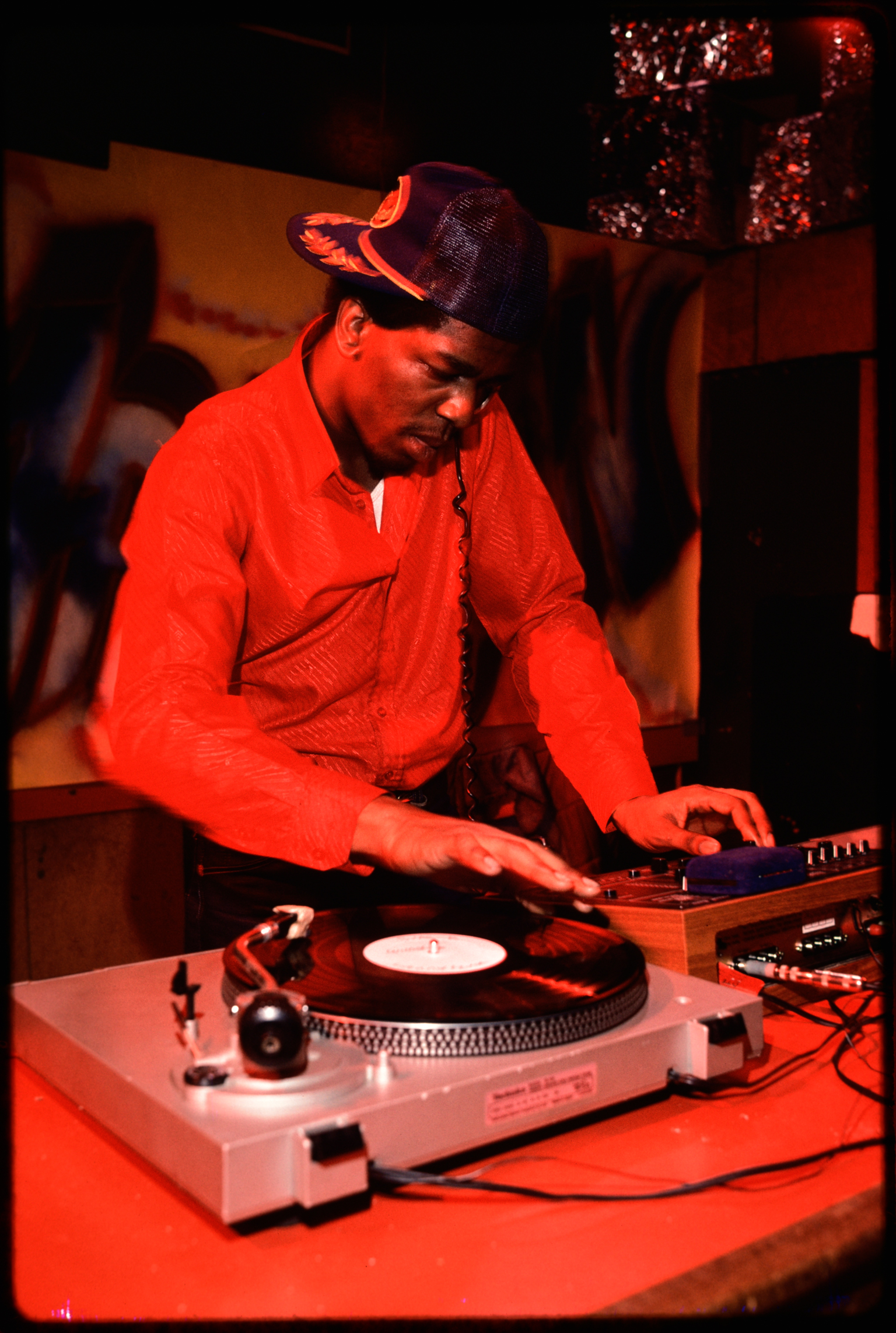 DJ wearing cap and red shirt operating turntables and mixing equipment, bathed in red stage lighting with wooden backdrop.
