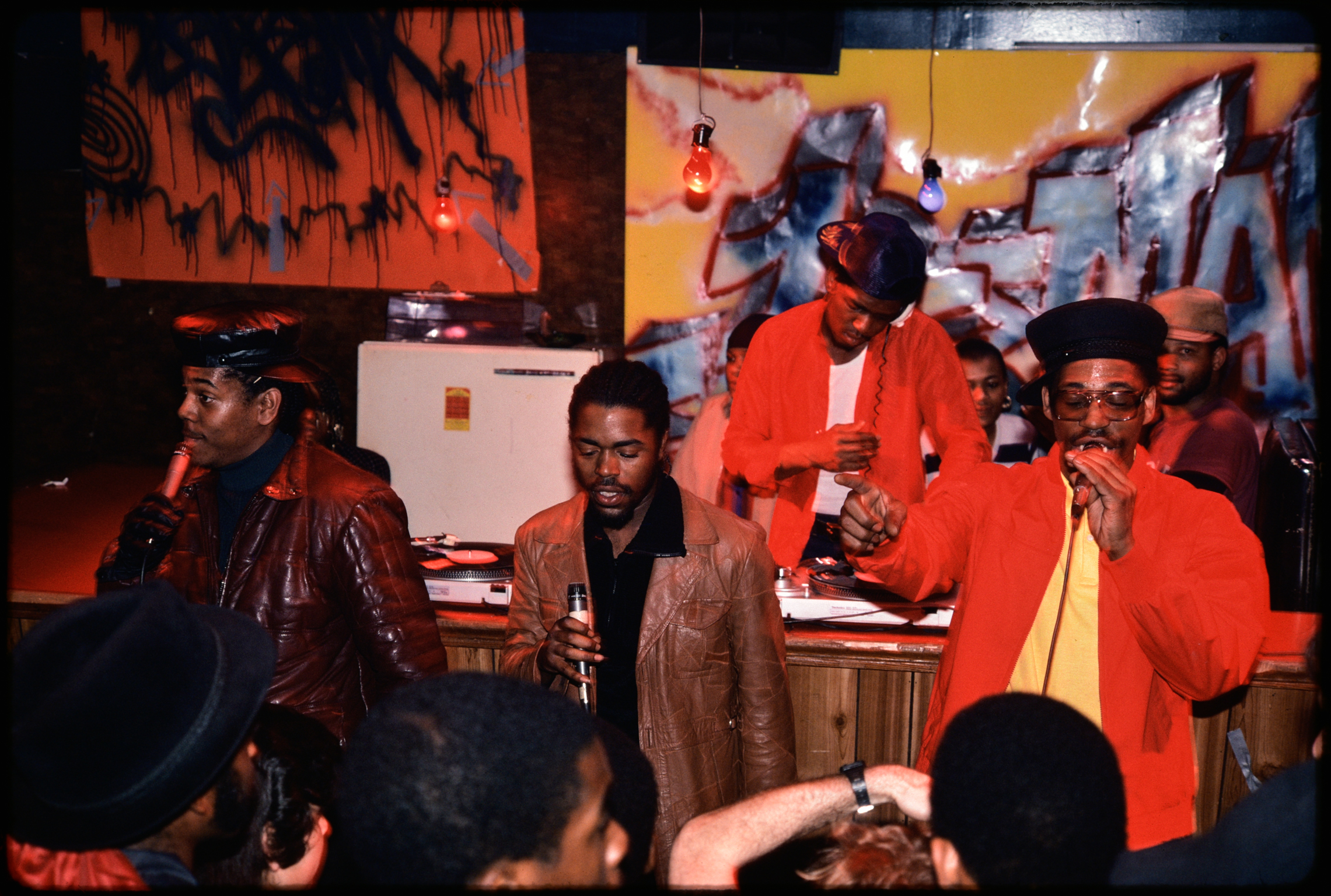 Five men in colourful clothing seated and standing around wooden table, with red and orange artwork on walls behind them.