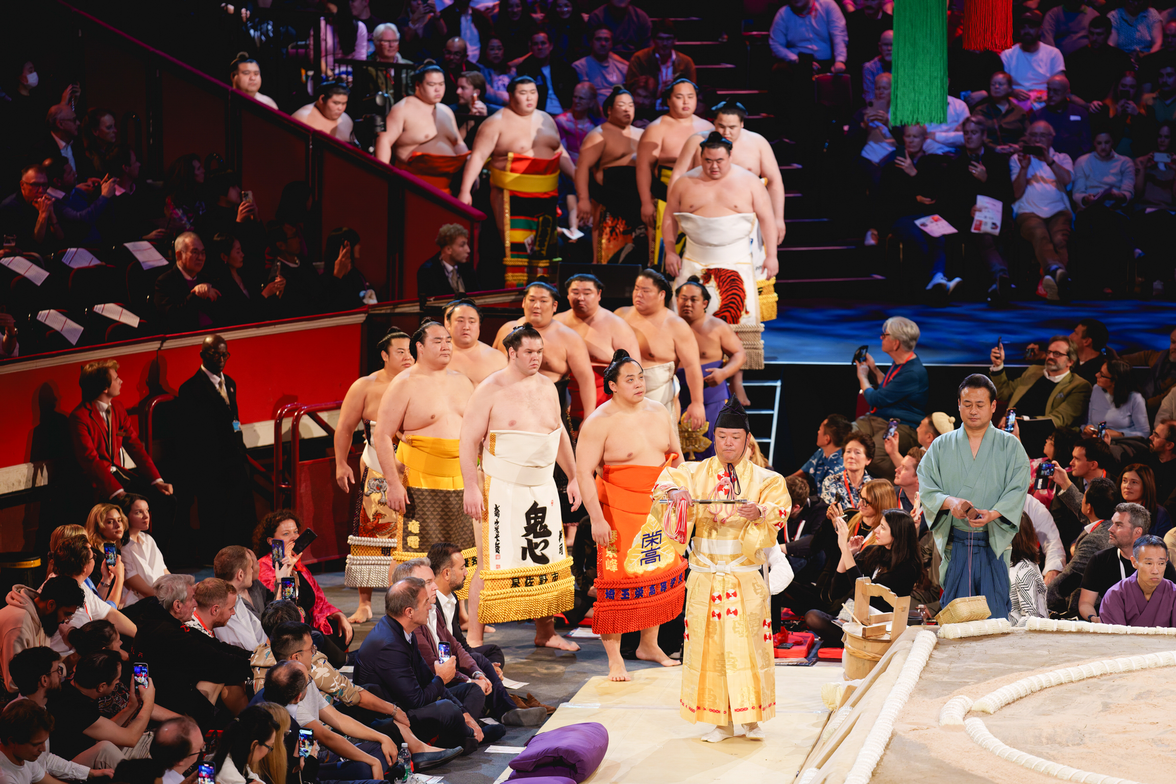 Group of sumo wrestlers in colourful mawashi standing on raised platform, surrounded by seated spectators in arena setting.
