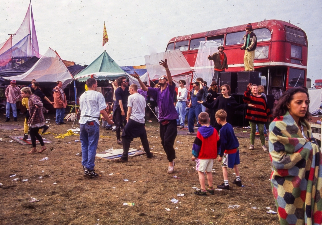 Crowded festival site with tents, stalls and an illuminated red double-decker bus. Groups of people, including children, milling about on the muddy ground.