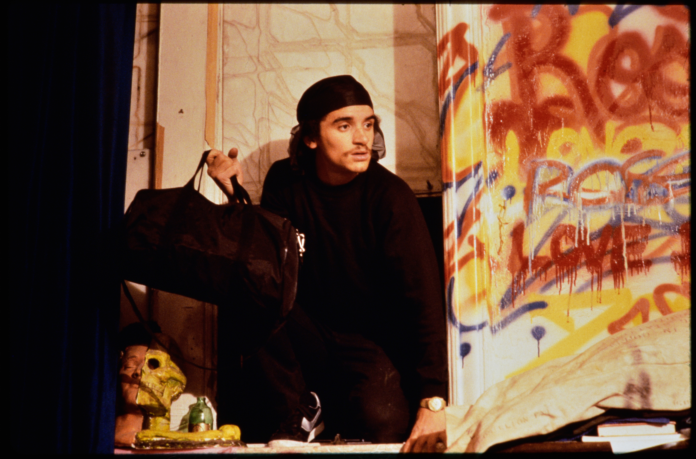 Person in black clothing and beanie stands in art studio with colourful graffiti-style paintings featuring orange, yellow, and blue tones.