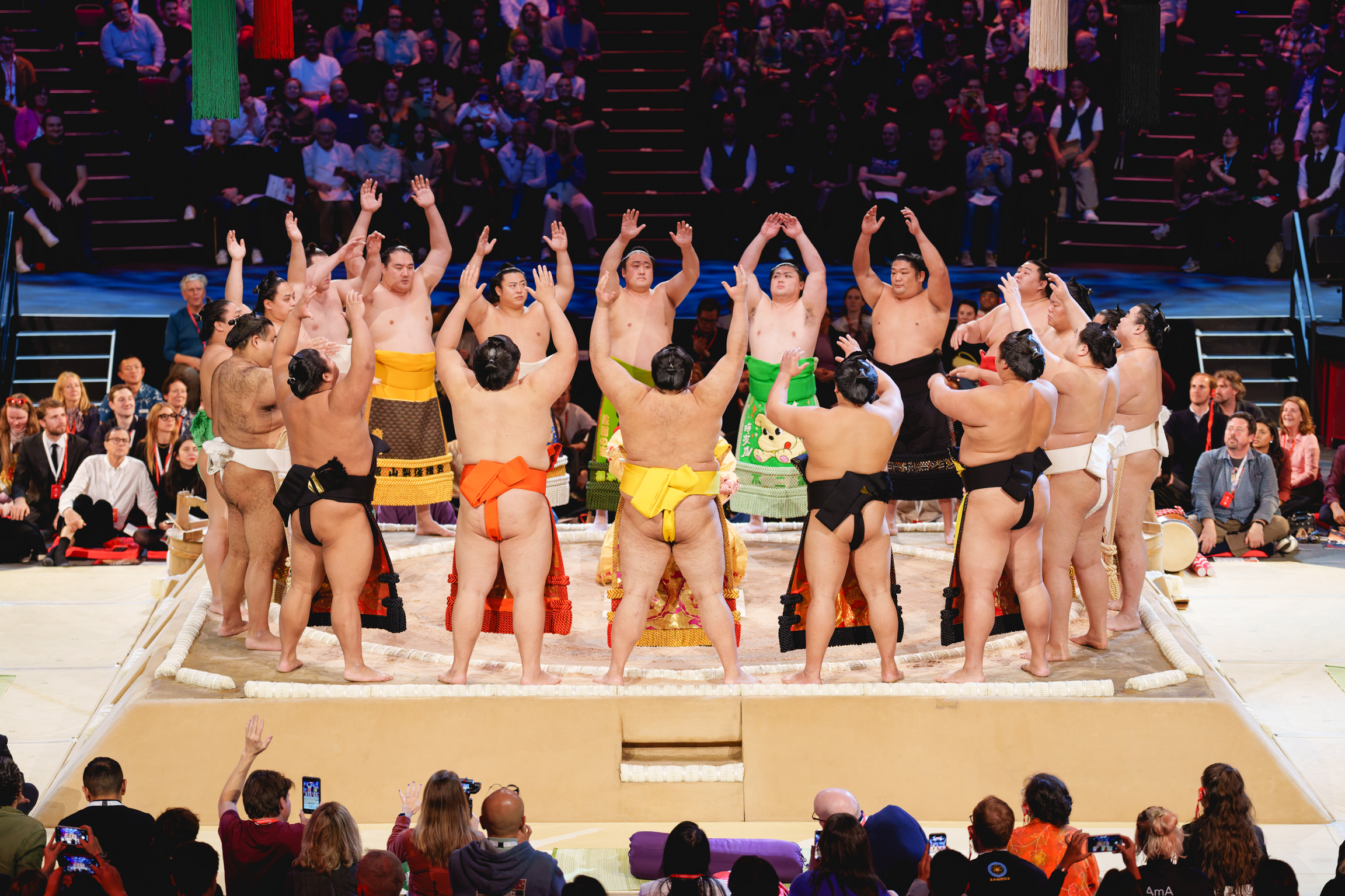 Sumo wrestlers in colourful mawashi belts standing in circle with raised arms on wrestling ring, crowd watching from stadium seating.