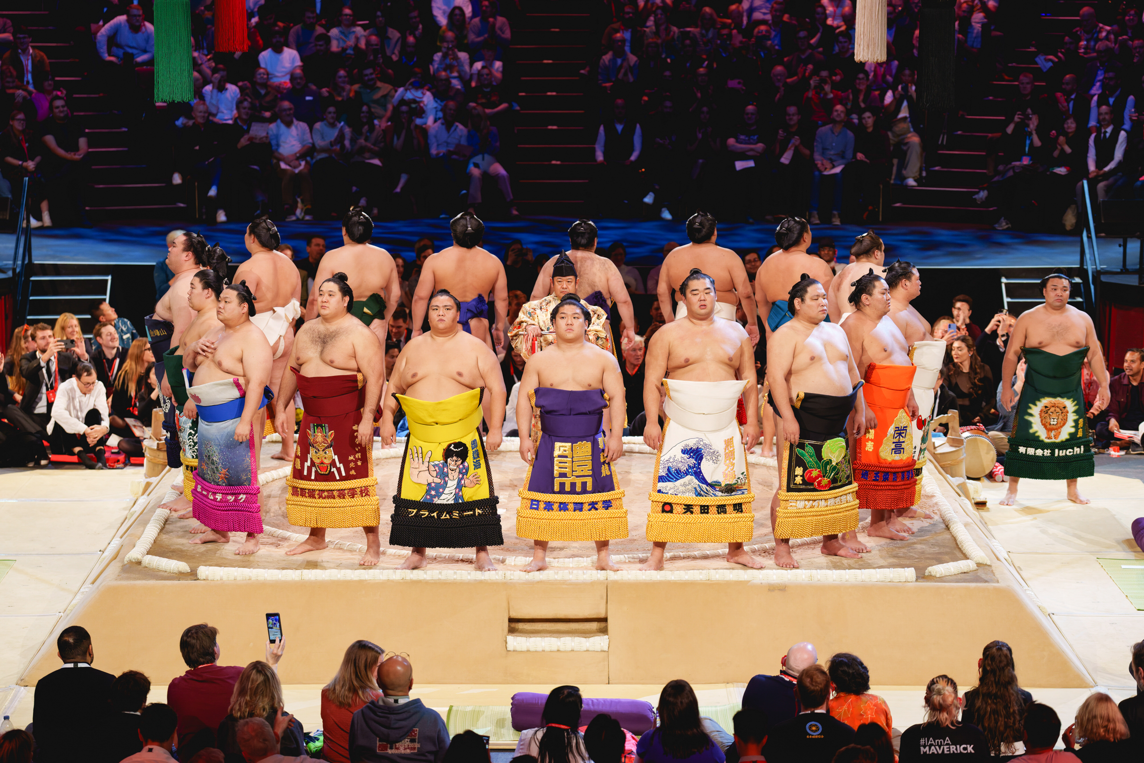 Sumo wrestlers in colourful ceremonial aprons lined up on sandy arena floor, surrounded by tiered seating filled with spectators.