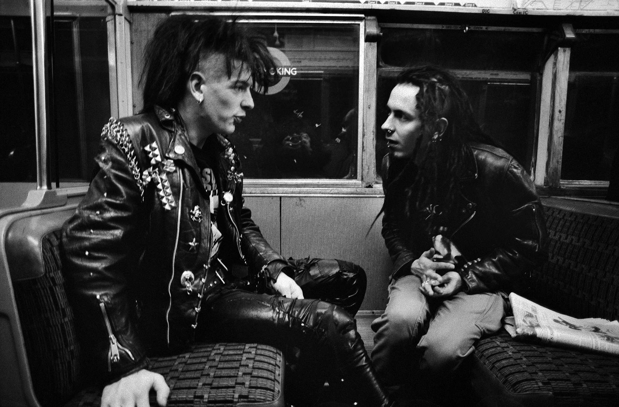 Two people with dark hair in leather jackets sitting on public transport seats in black and white photograph.