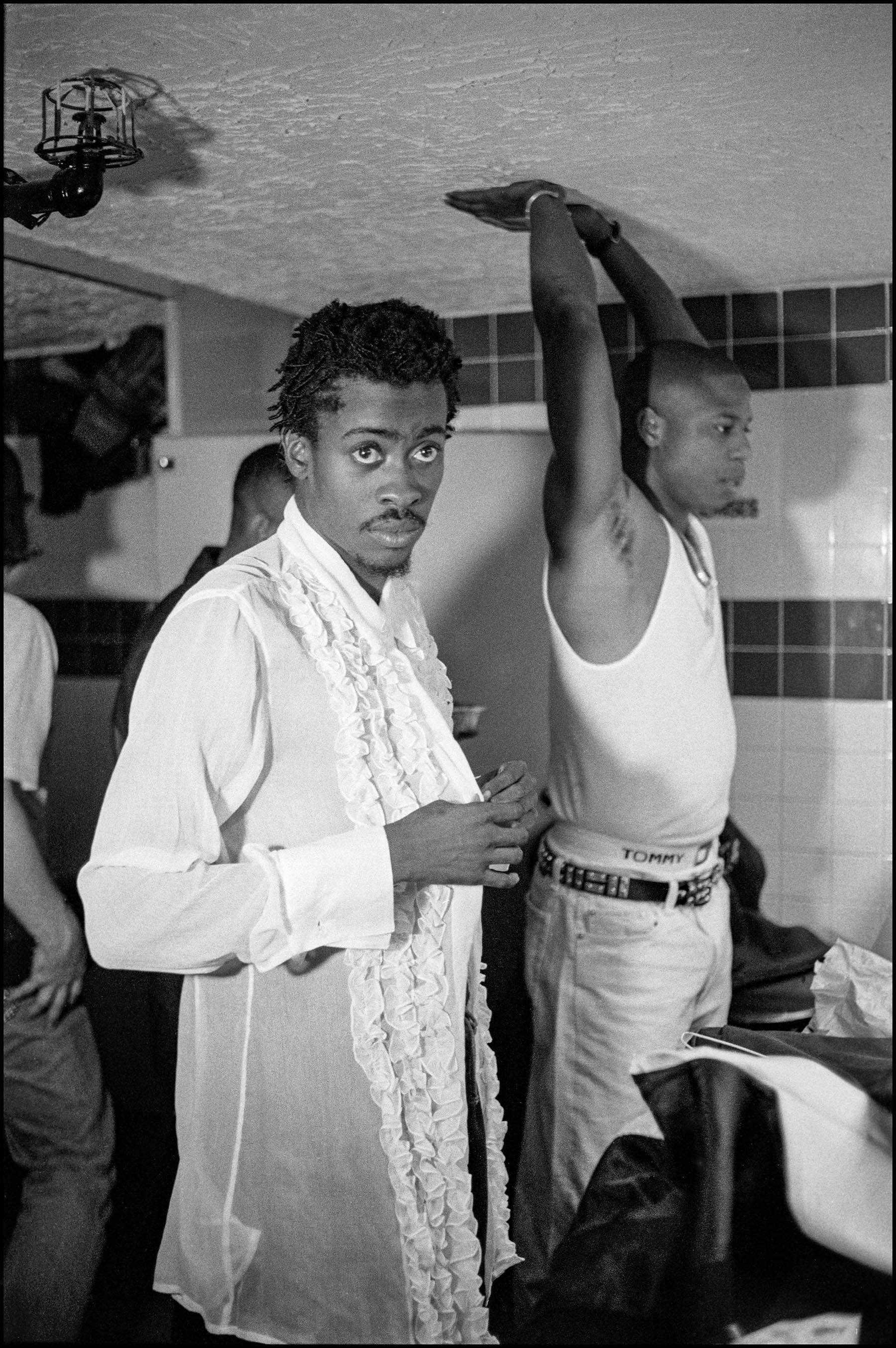 Two young men, one wearing a white ruffled shirt and the other wearing a white tank top, standing together in a room with windows in the background.
