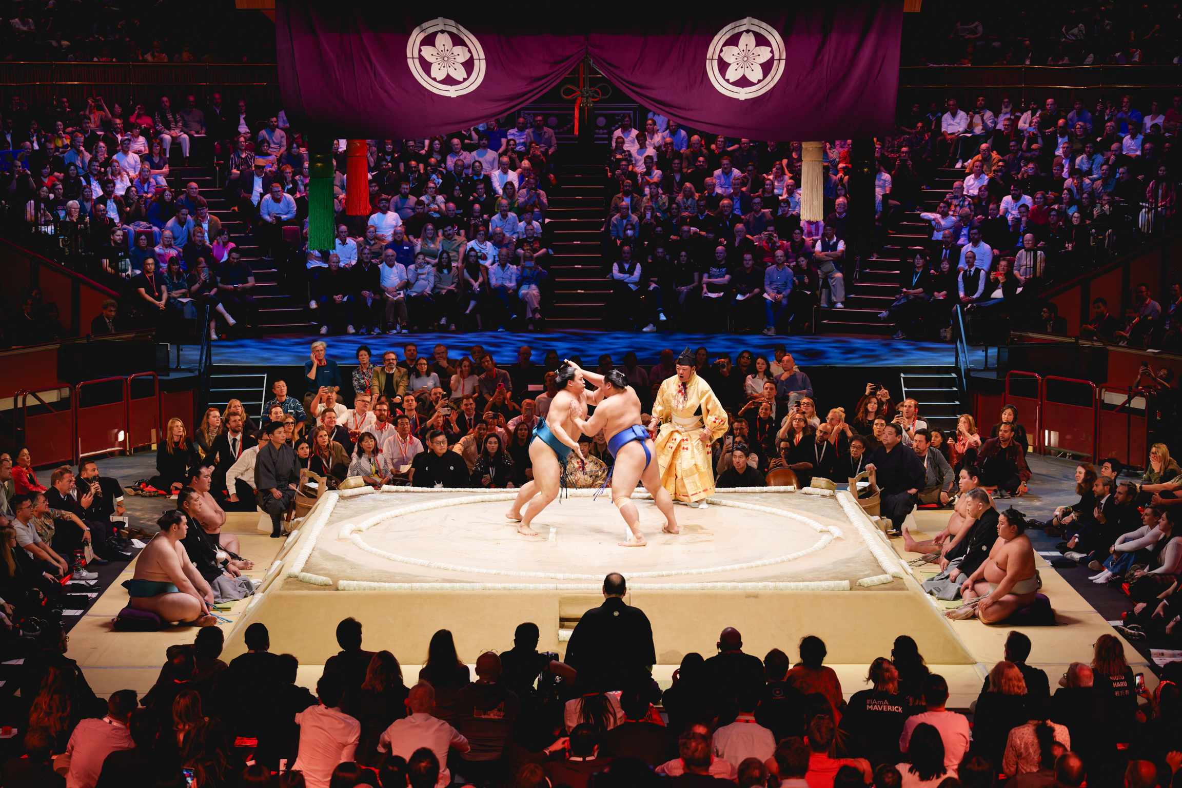 Sumo wrestlers competing on white mat in arena surrounded by seated spectators, with purple banners displaying circular emblems overhead.