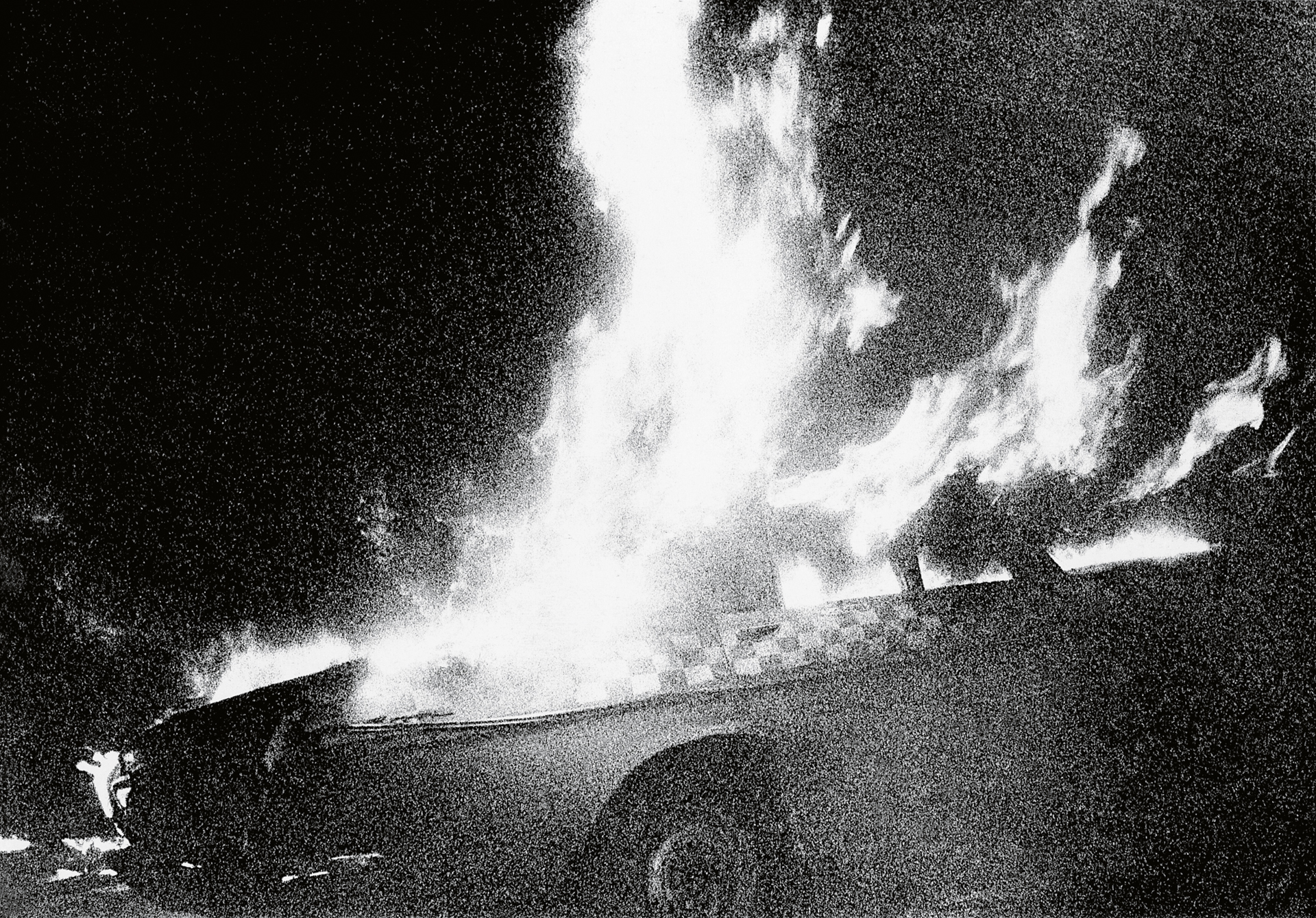 Black and white image showing bright white flames and smoke rising against dark background, with grainy texture throughout.