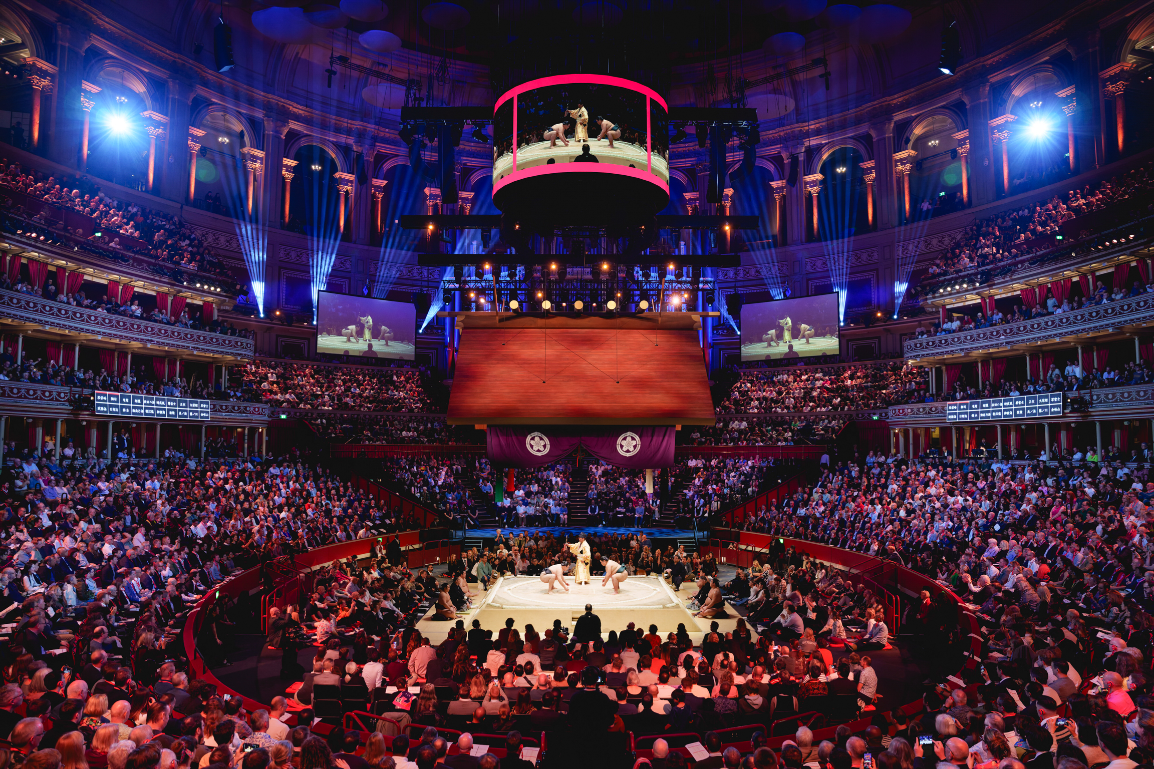 Interior of Royal Albert Hall during performance with packed circular seating tiers, red upholstery, ornate ceiling, and illuminated stage.
