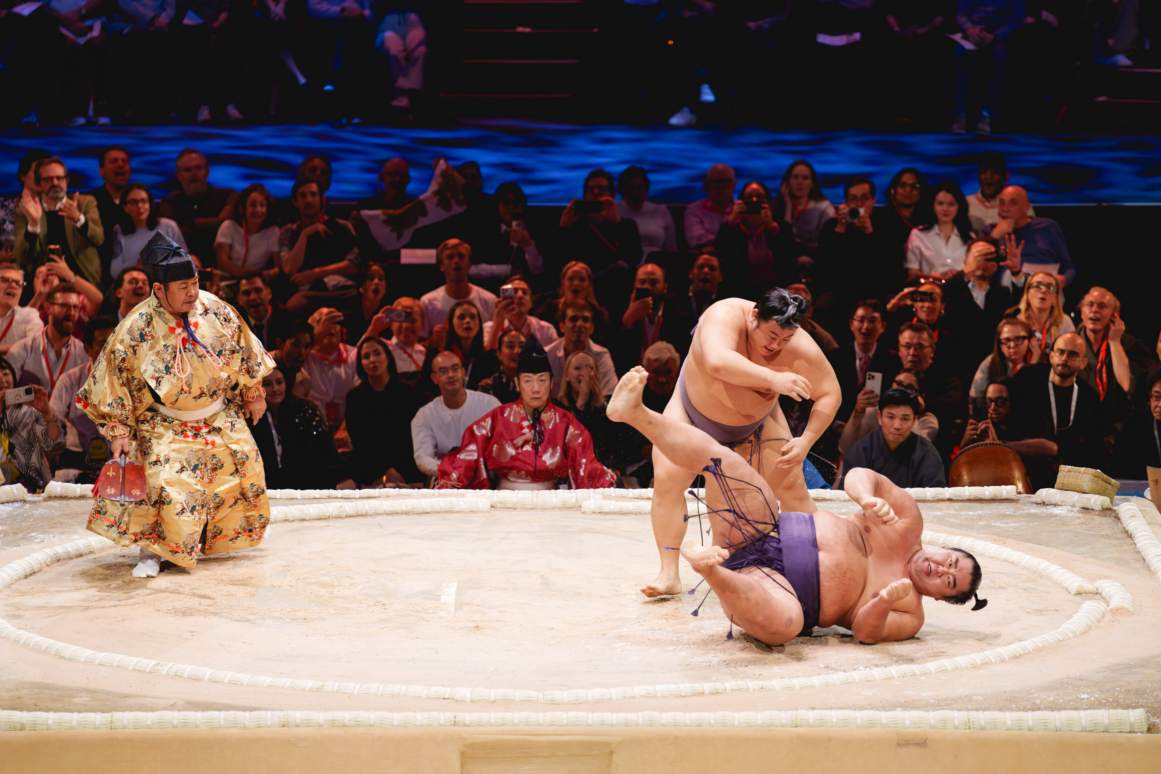 Sumo wrestlers grappling on circular mat with referee in patterned robe nearby and crowd of spectators in tiered seating background.