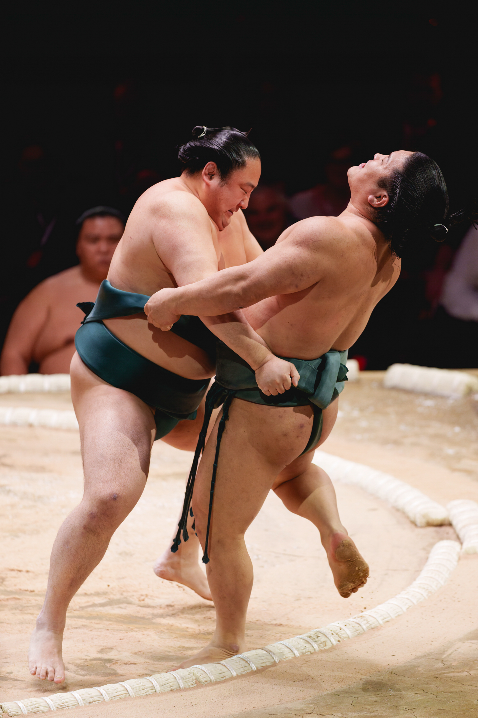 Two sumo wrestlers grappling in a ring, wearing traditional mawashi belts, with spectators visible in blurred background.