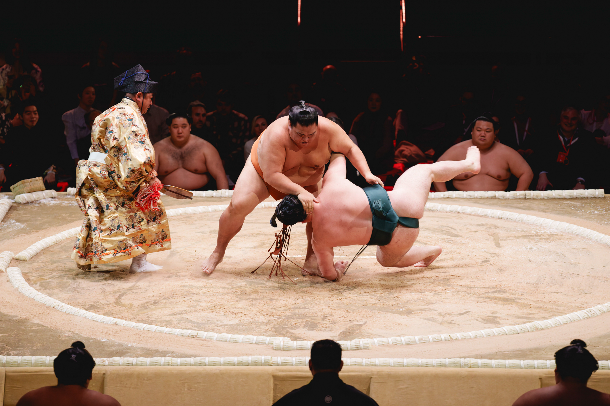 Two sumo wrestlers grappling in circular ring whilst referee in ornate robe observes. Audience visible in background and foreground.