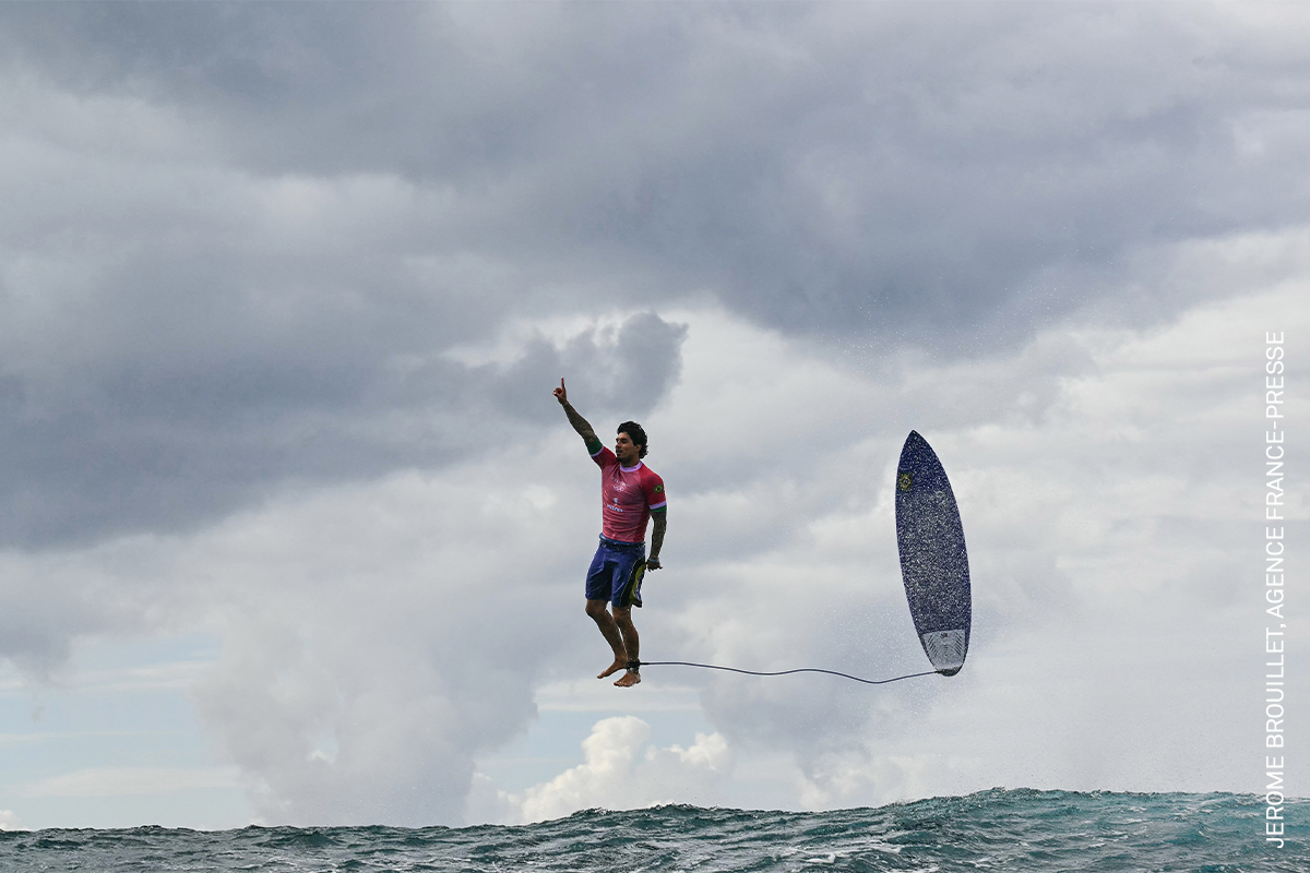 Surfer mid-jump, silhouetted against an overcast sky, with a surfboard on the water below.