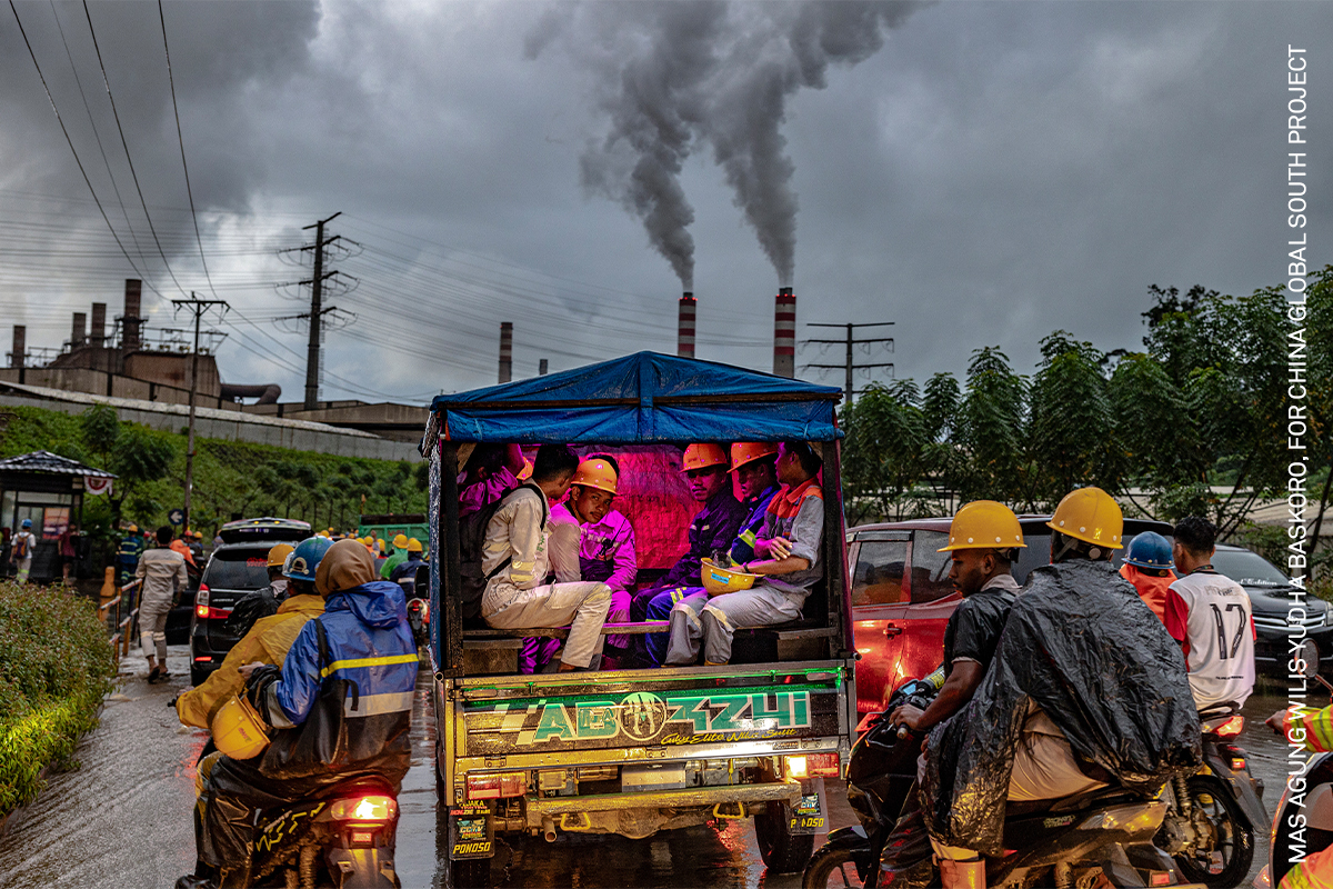 Colourful open-top truck filled with commuters on a rainy urban street, with industrial chimneys emitting smoke in the background.