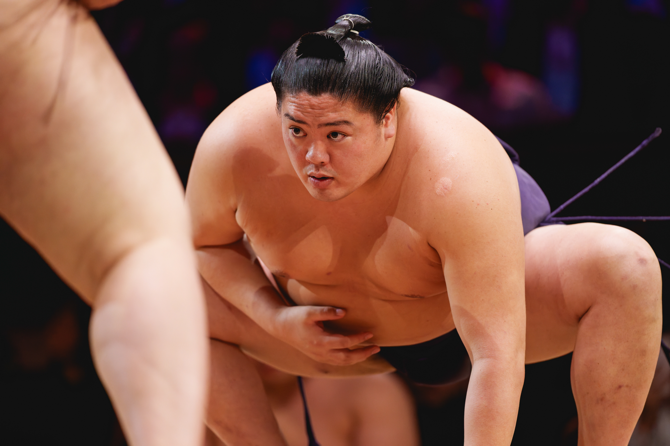 Two sumo wrestlers grappling in crouched position during match, with dark hair in traditional topknots against blurred purple background.