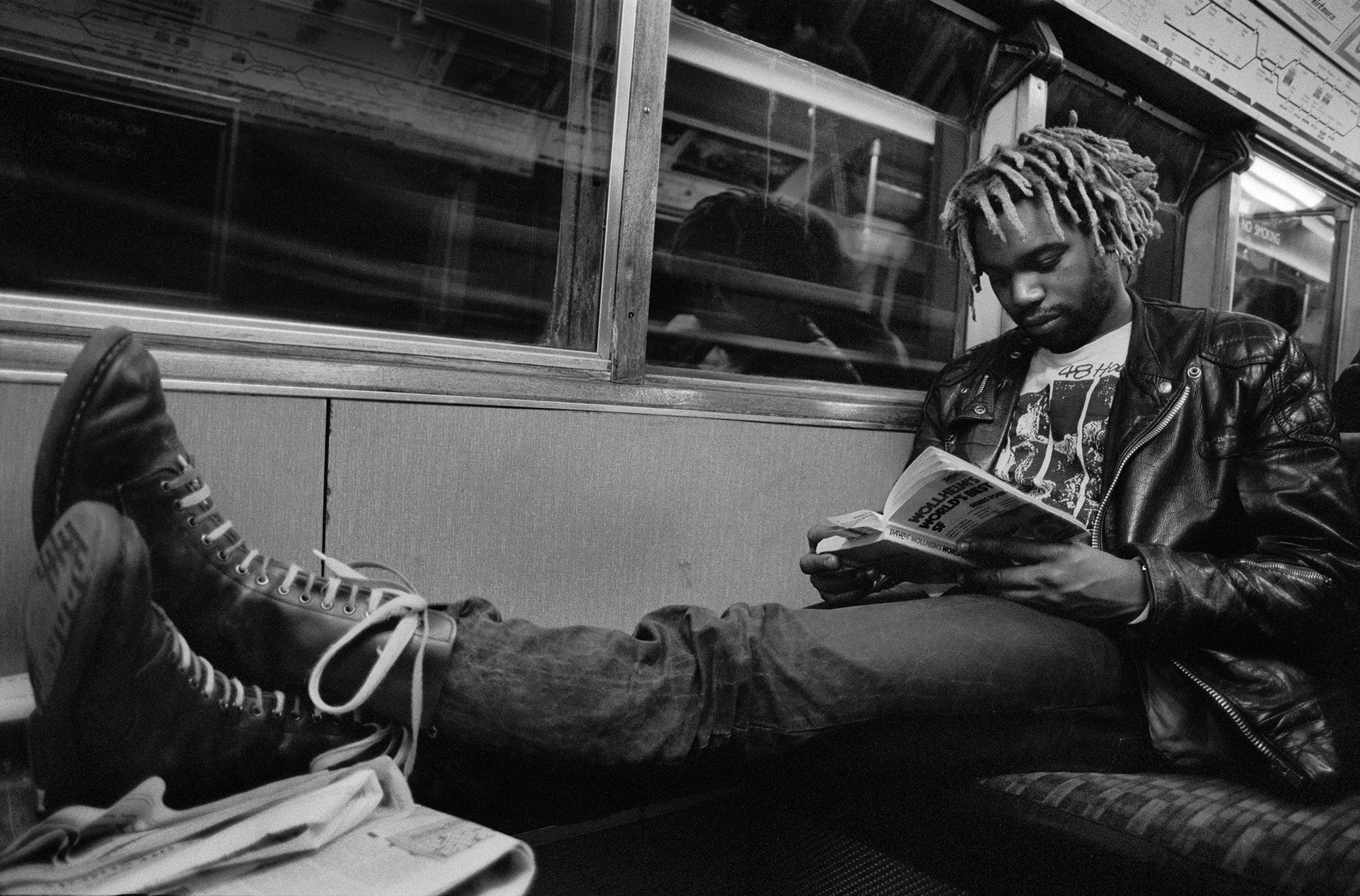 Person with dreadlocks reading book whilst sitting on bench inside what appears to be a train or bus carriage. Black and white image.
