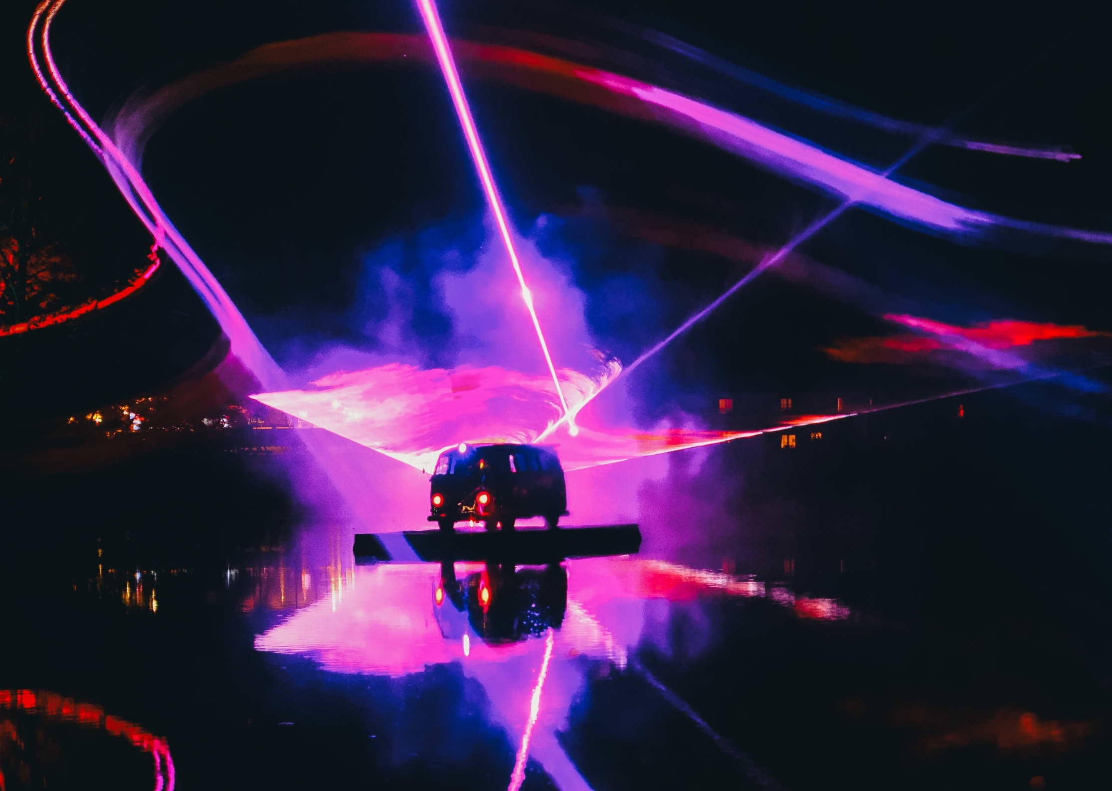 Bright pink and purple laser beams crossing over dark stage with silhouetted equipment, blue and magenta light trails against black background.