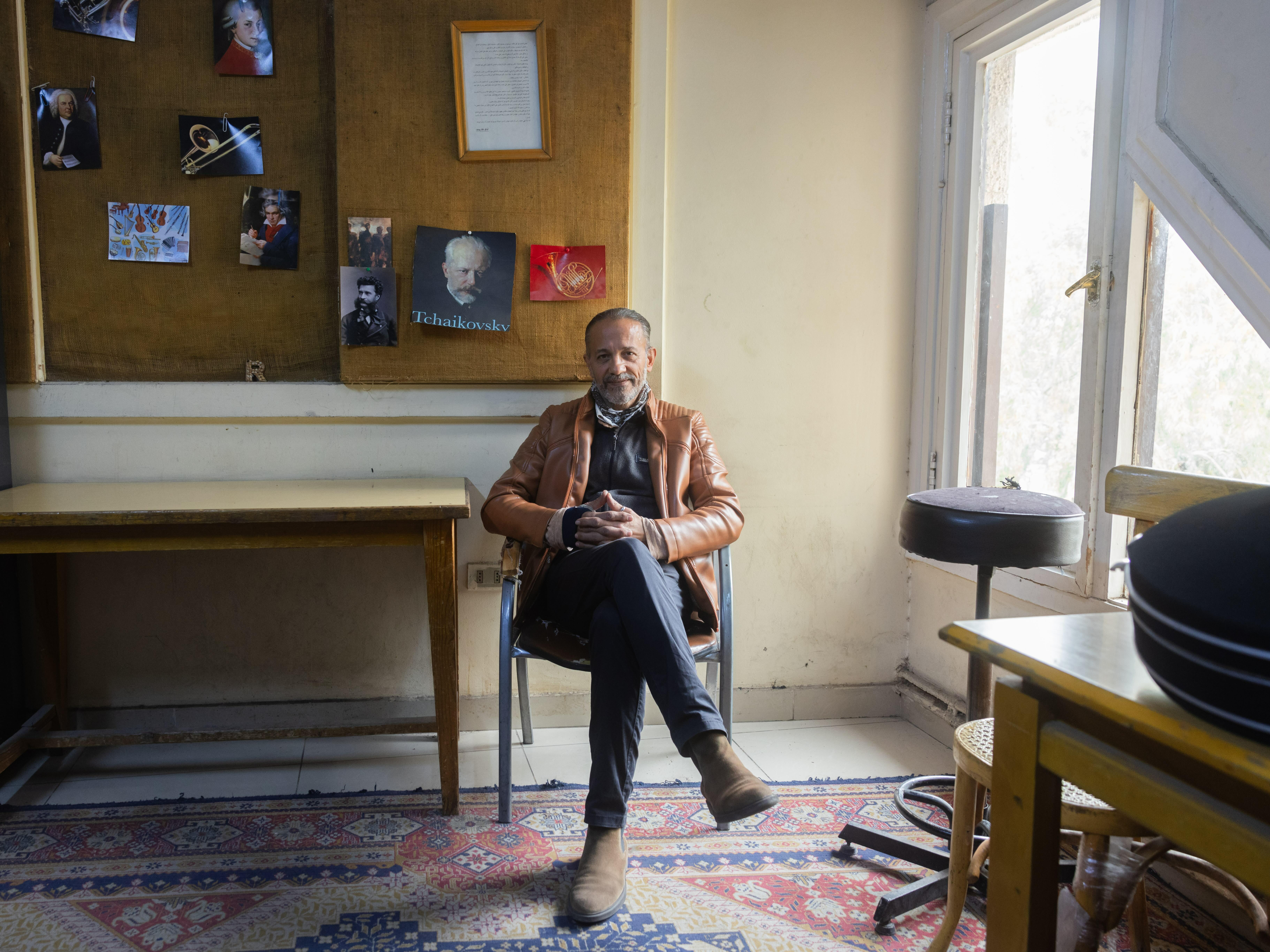 An elderly man sits in a room with framed artwork on the walls and various furnishings around him. He appears to be deep in thought, with a contemplative expression on his face.
