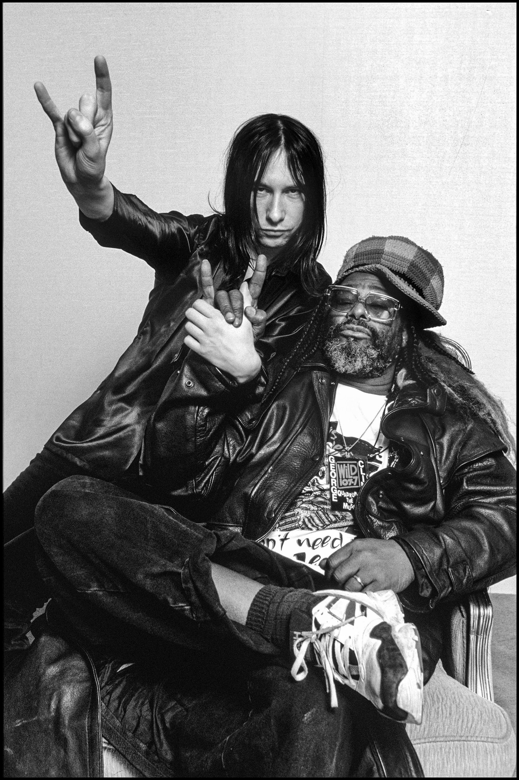 Black and white image of two men, one making a peace sign with his hand and the other wearing a cap and leather jacket.