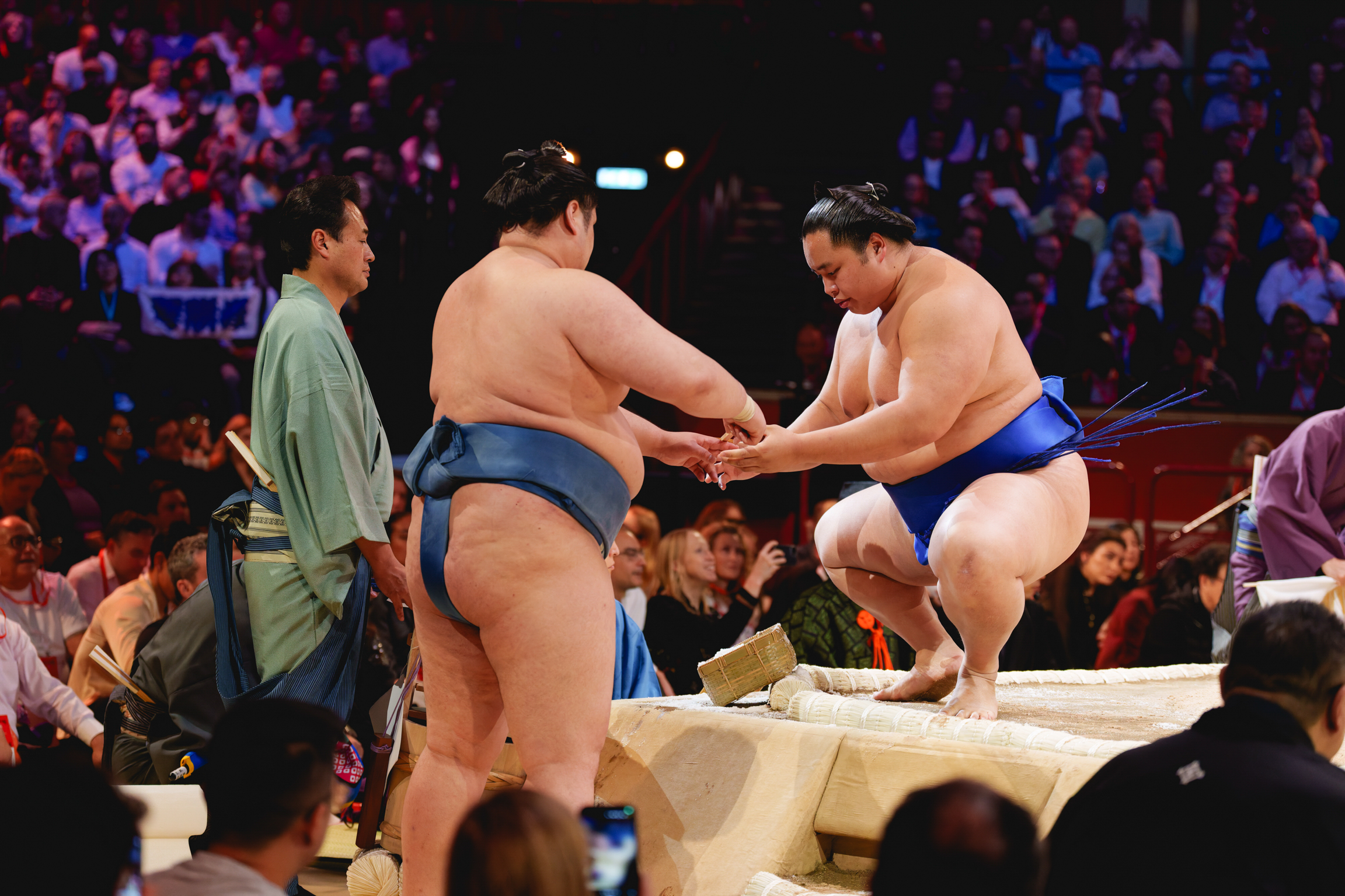 Two sumo wrestlers grappling in centre of ring with referee in green robes nearby, surrounded by seated spectators in arena.