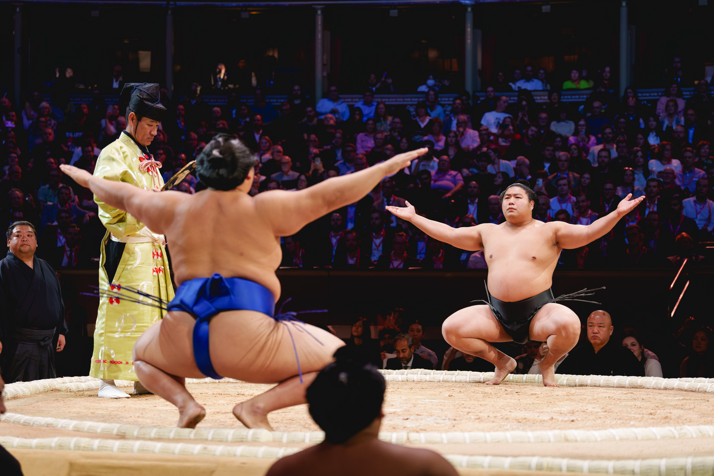 Two sumo wrestlers in blue mawashi on sandy ring, referee in yellow robes between them, crowded stadium in background
