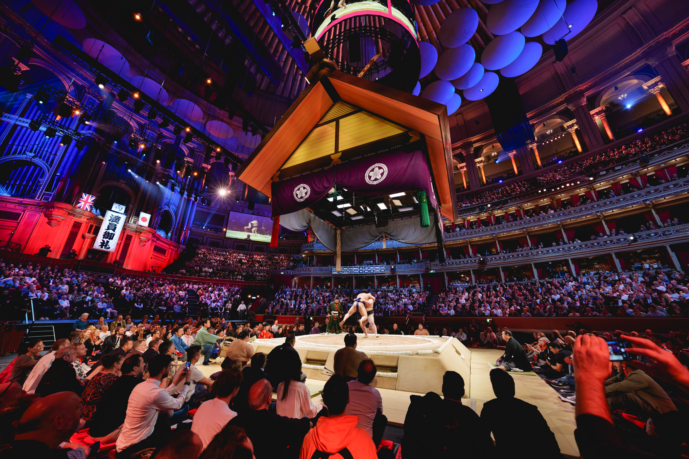 Large arena with central stage, audience in tiered seating, suspended orange and purple geometric structures overhead, dramatic lighting.