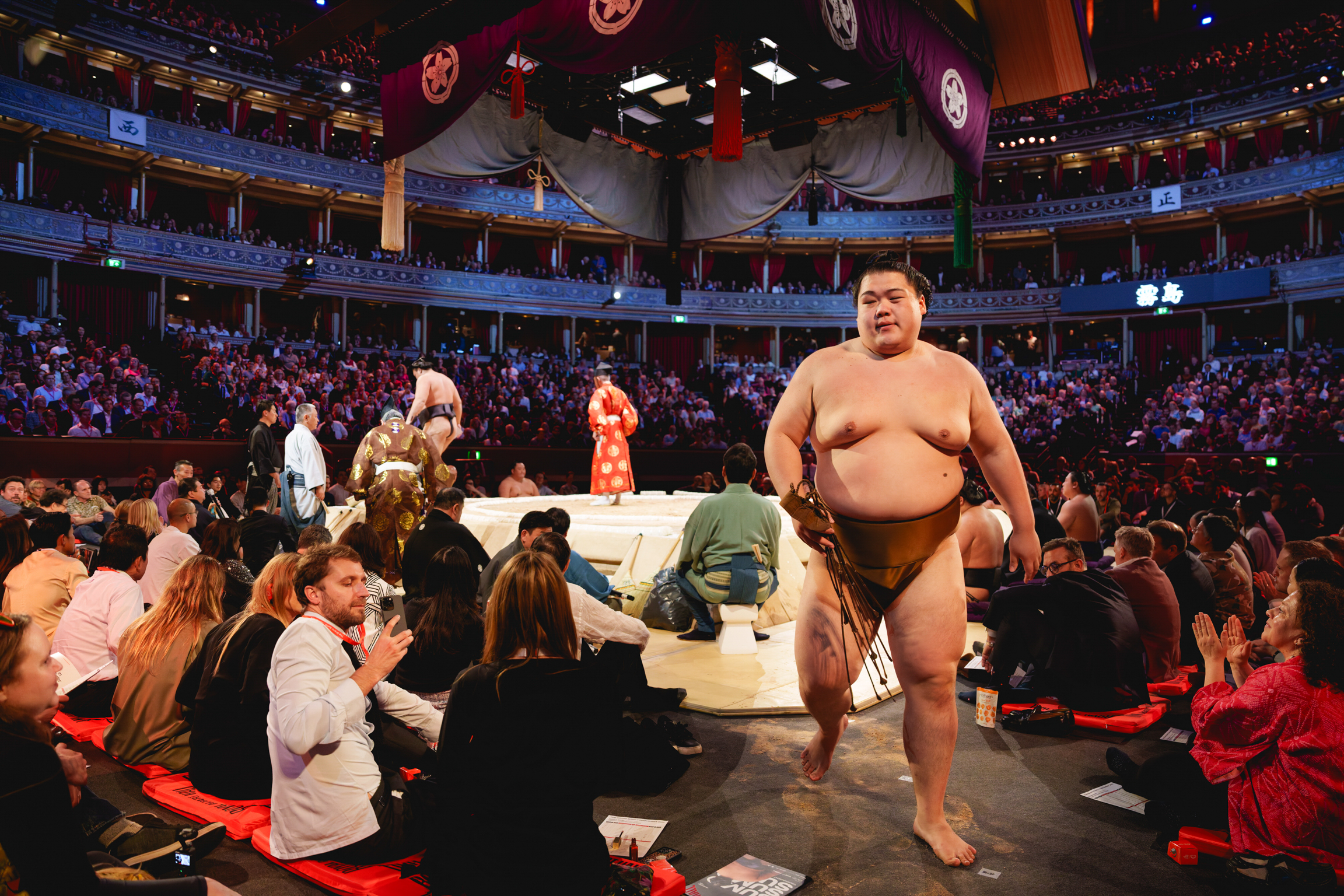 Sumo wrestler in green belt stands in arena whilst spectators sit on red cushions around white wrestling ring.