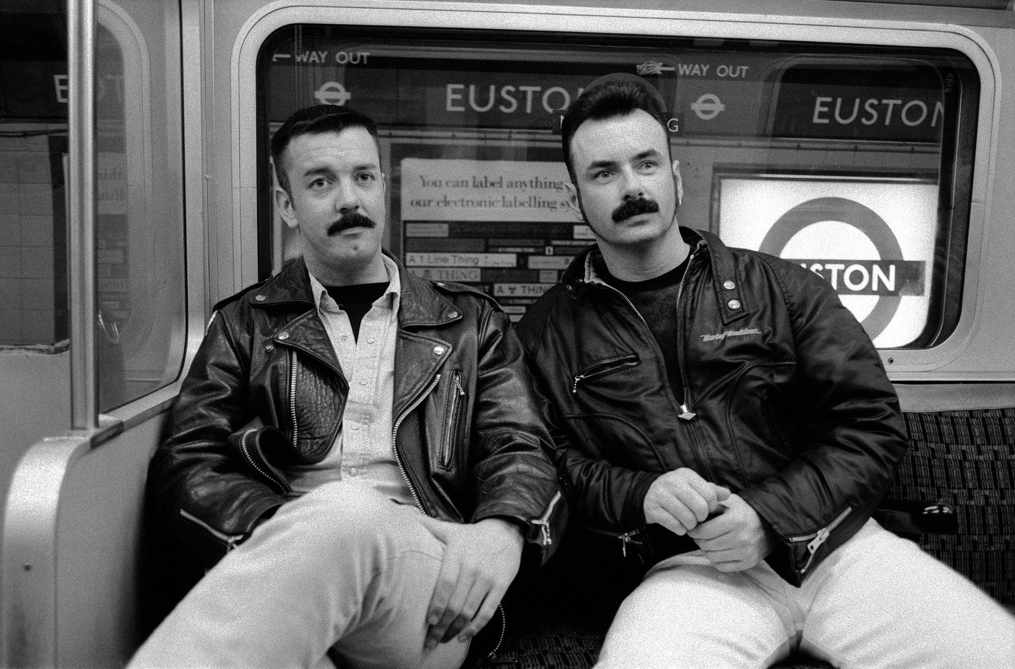Two men with moustaches wearing leather jackets sitting on train seats, black and white image with "EUSTON" visible on windows.