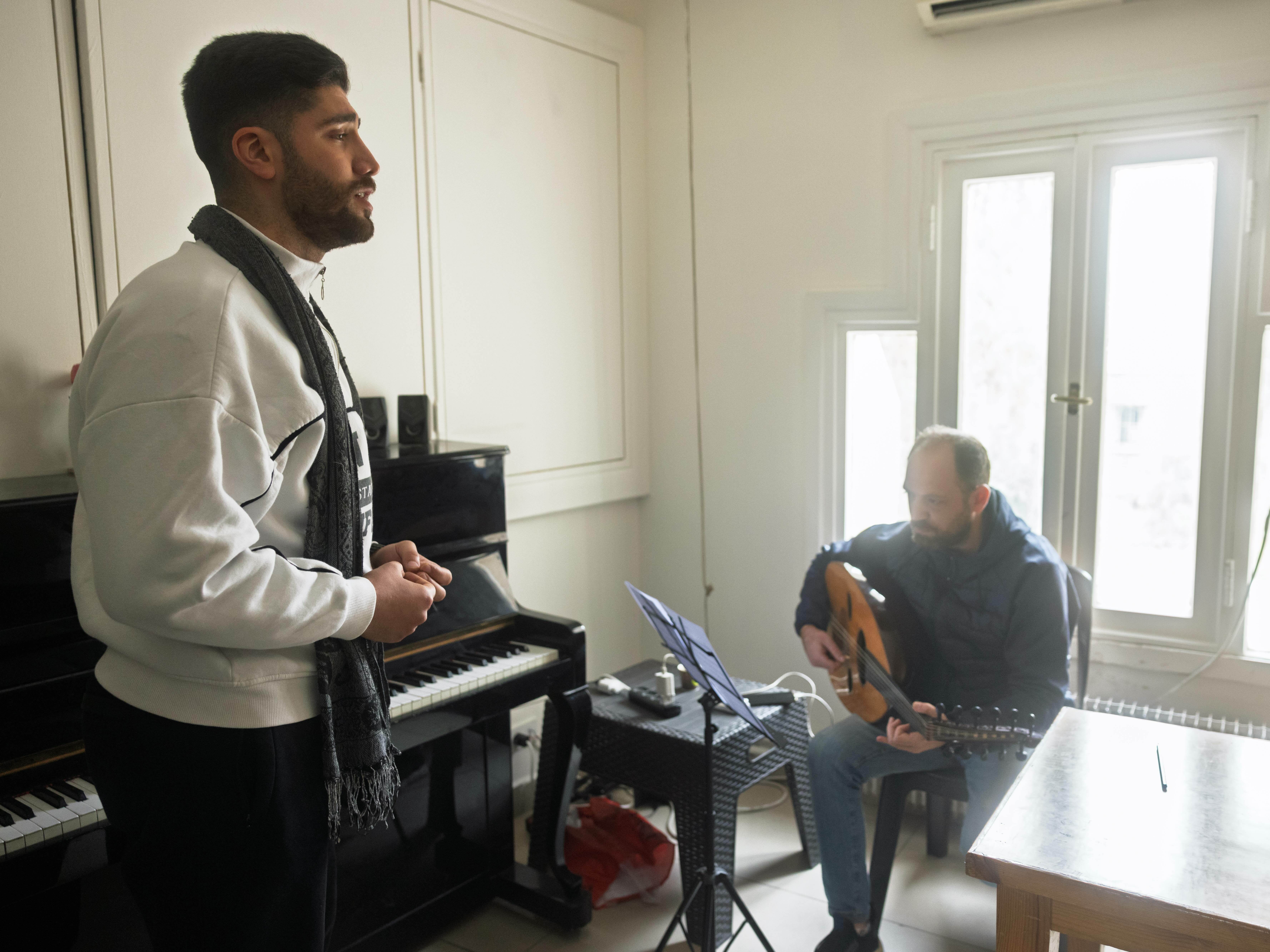 A man with a beard standing near a piano, a second man seated in a chair holding a guitar.