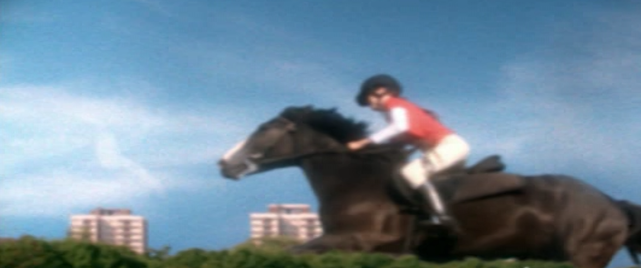 A person riding a brown horse at a racetrack, with tall buildings in the background.